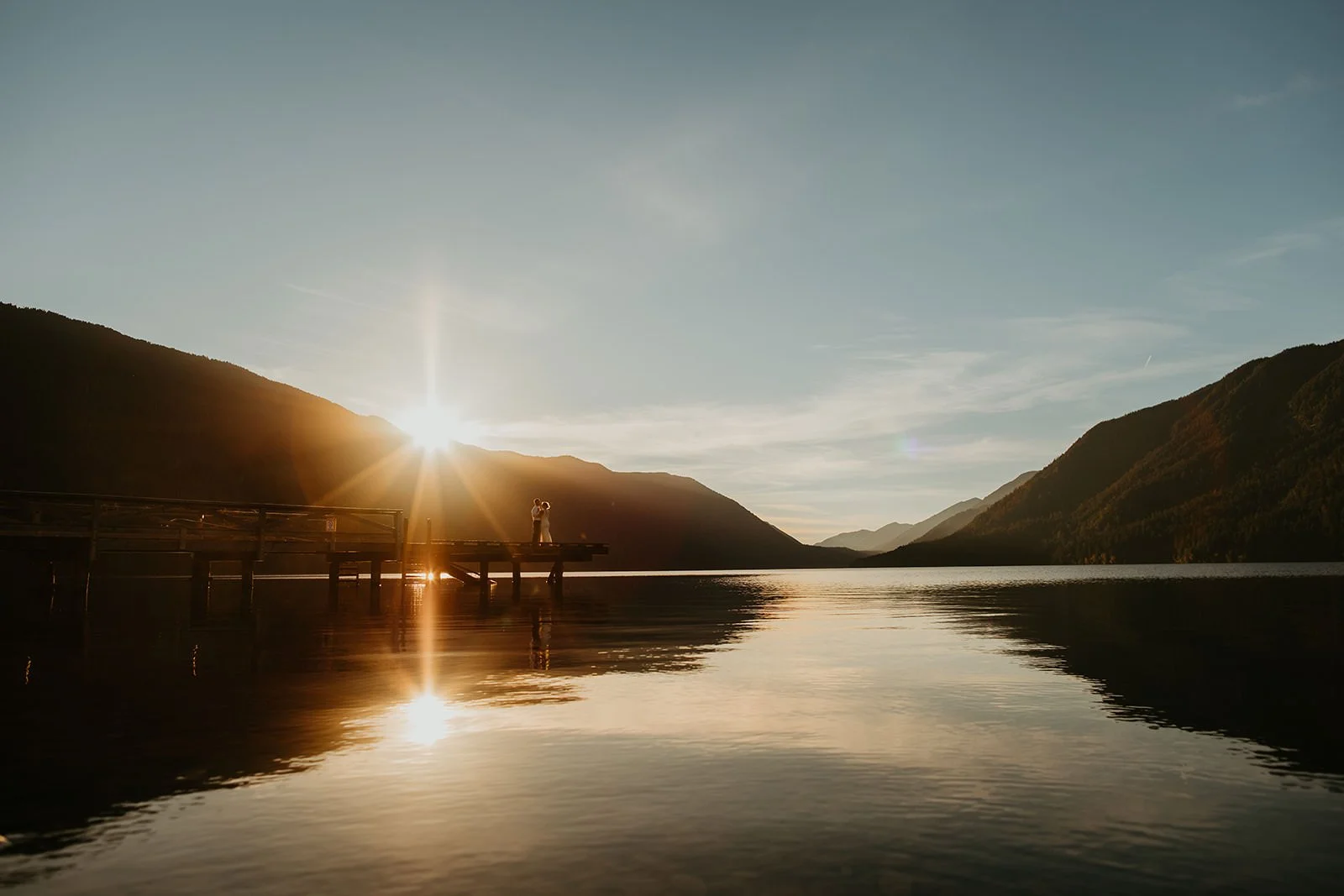 Couple standing at the edge of a wooden dock on a calm lake at sunset, surrounded by forested hills reflected in the water.