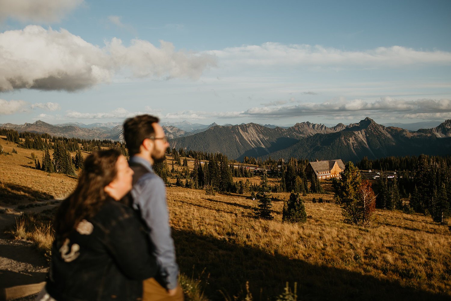Couple standing side by side overlooking a wide mountain valley with layered peaks in the distance.