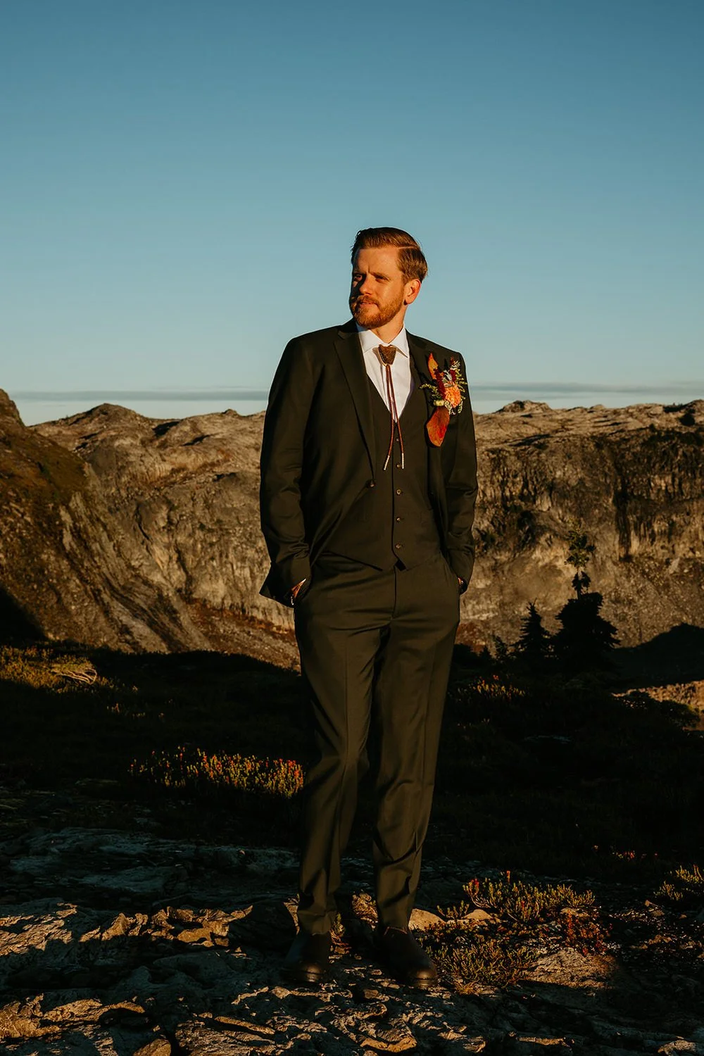 A person stands alone on a rocky mountain ridge in warm evening light, with expansive alpine terrain and distant peaks in the background.