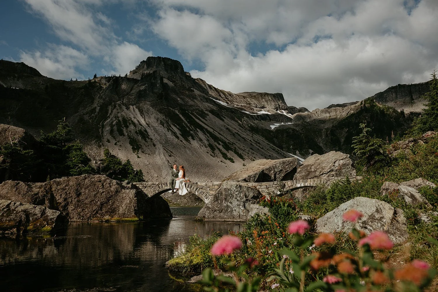 Eloping couple sitting on a small stone bridge over a mountain lake, surrounded by rocky terrain and alpine scenery