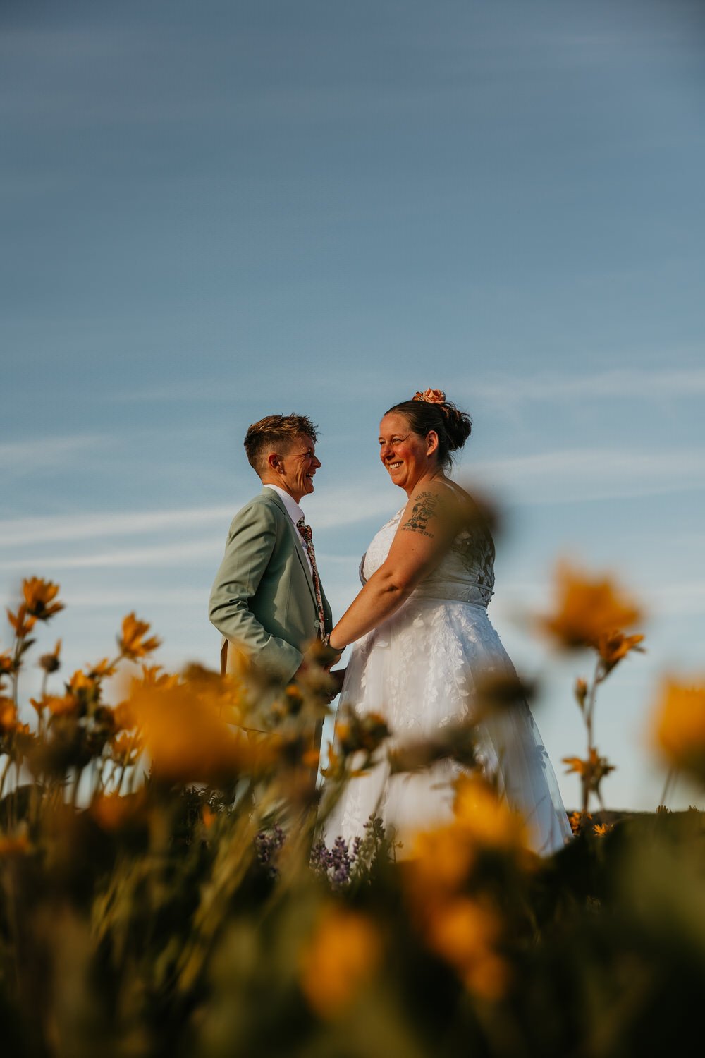 Two brides holding hands and smiling at each other in a field of wildflowers under a clear blue sky during a summer elopement.