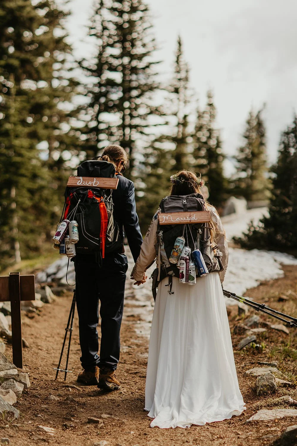 Newly married couple hiking hand in hand on a forest trail wearing backpacks labeled just married, with snow patches and evergreen trees around them