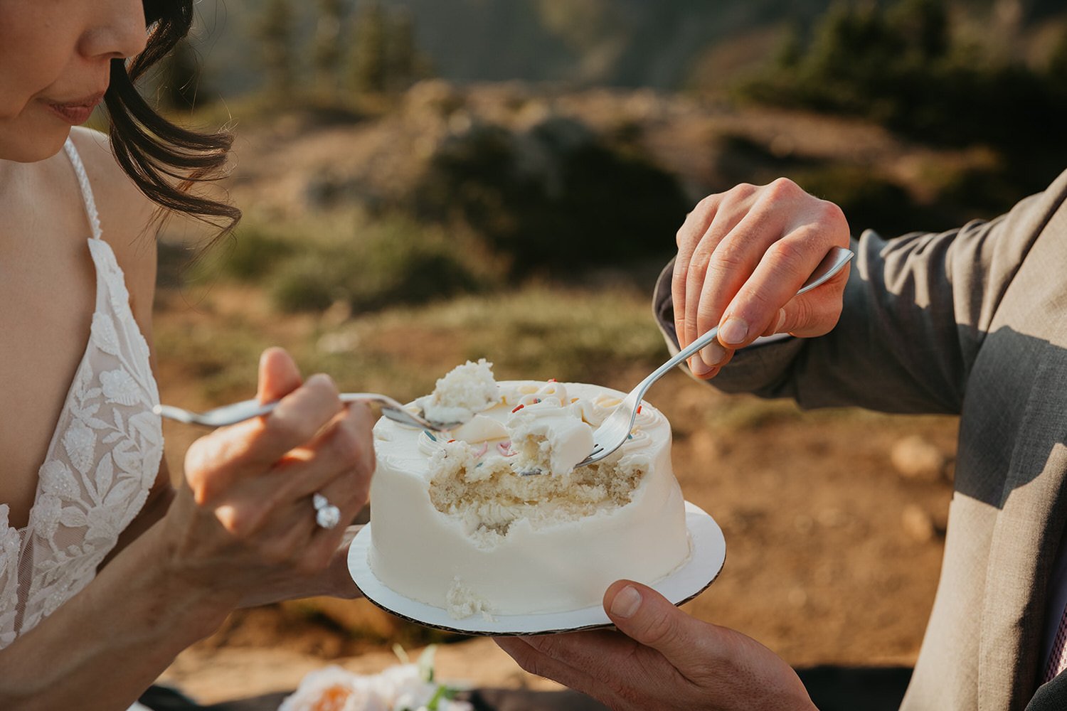 Close-up of hands sharing a small white cake outdoors, forks digging into the frosting.