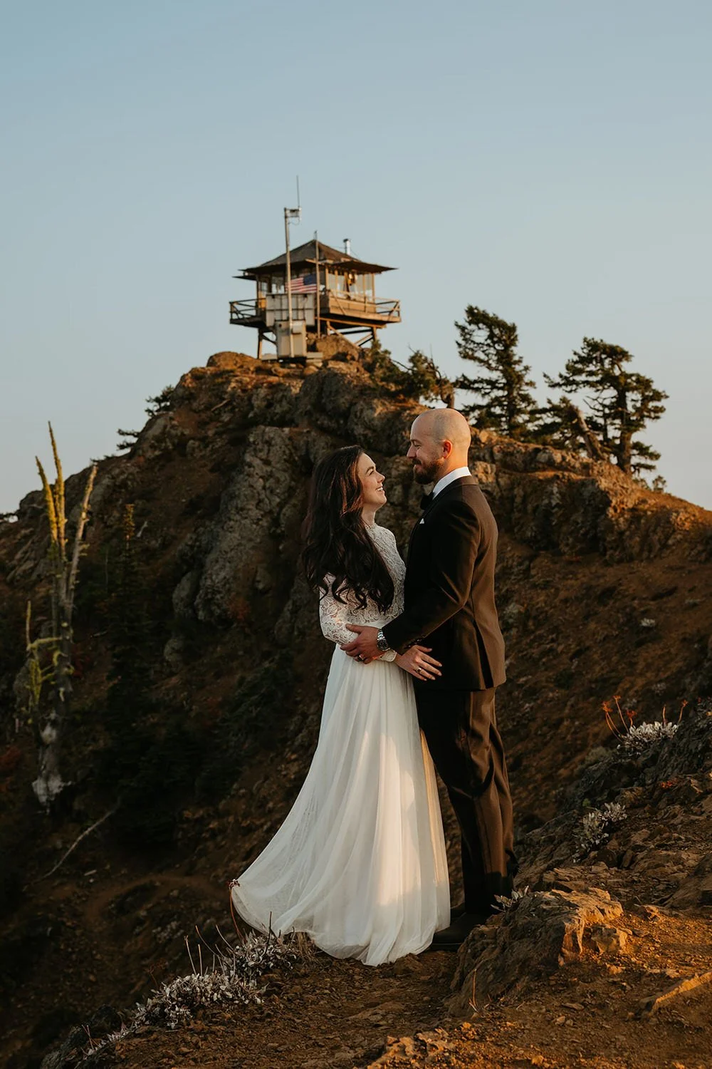 Couple standing together on a mountain overlook with a fire lookout in the background during a Washington elopement