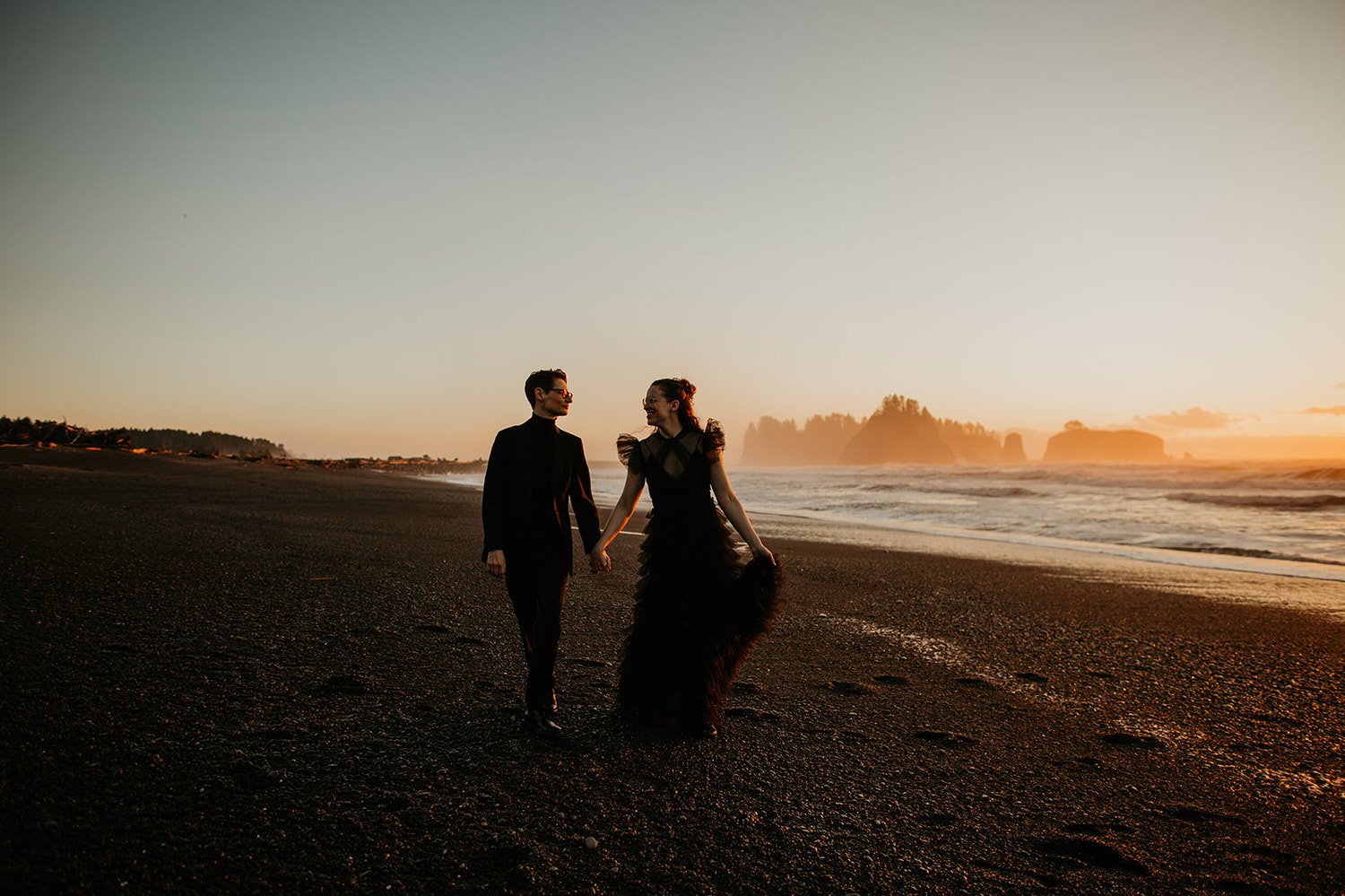 Eloping couple walking along a black sand beach at sunset on the Washington coast with sea stacks and waves behind them.