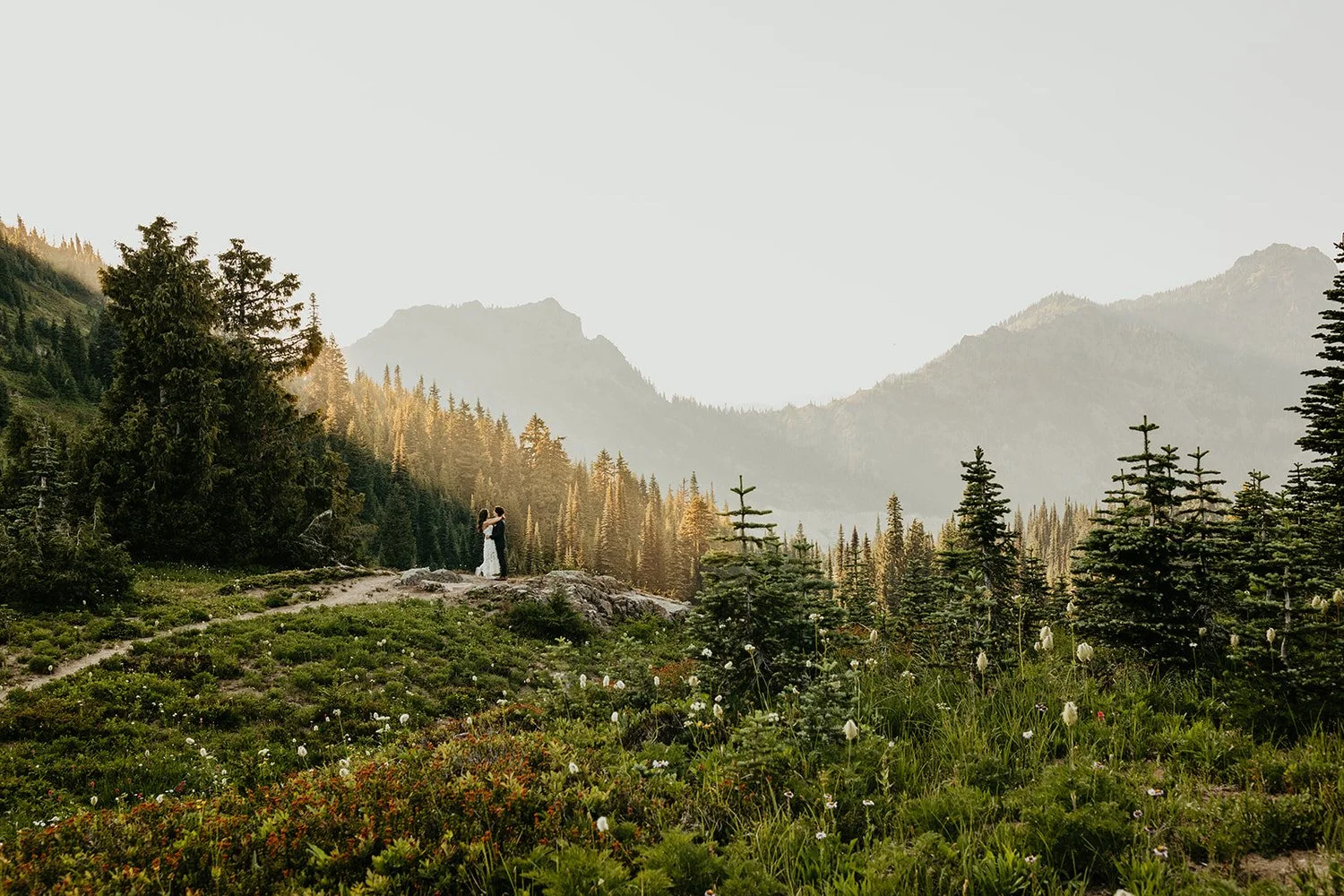 Couple standing on trail in high alpine meadow with white wildflowers scattered throughout; evergreen forest and misty dramatic peaks behind