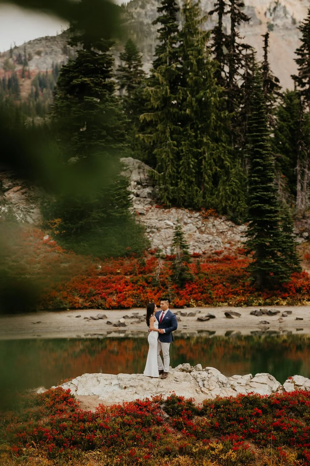A couple stands together on a small rock beside a calm alpine lake, surrounded by evergreen trees and fall-colored shrubs reflected in the water.