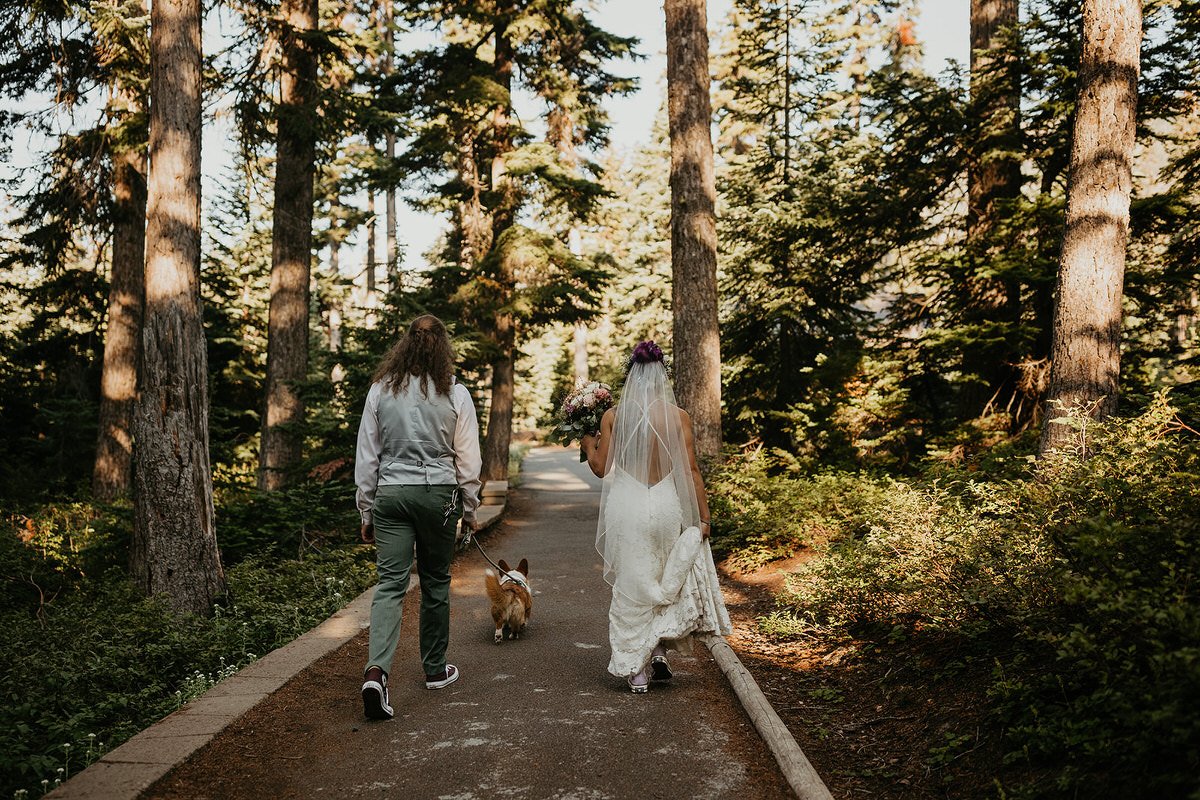 Couple walking hand in hand down a forest path after their ceremony, the bride holding her bouquet and dress while their small dog trots between them.