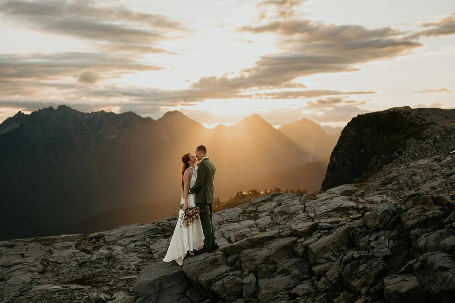 Couple sharing a kiss on a rocky summit as dramatic sunbeams break through clouds at sunset