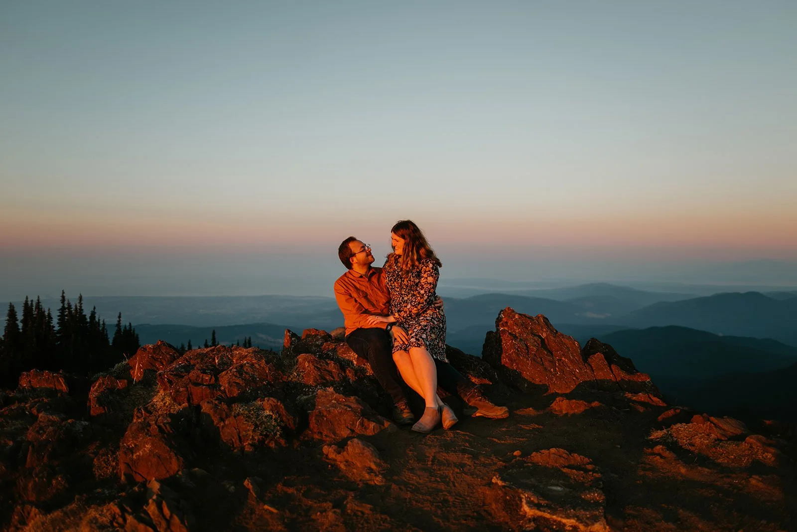 Couple sitting together on a rocky mountain summit at sunset, wrapped in warm golden light with layered hills fading into the distance.