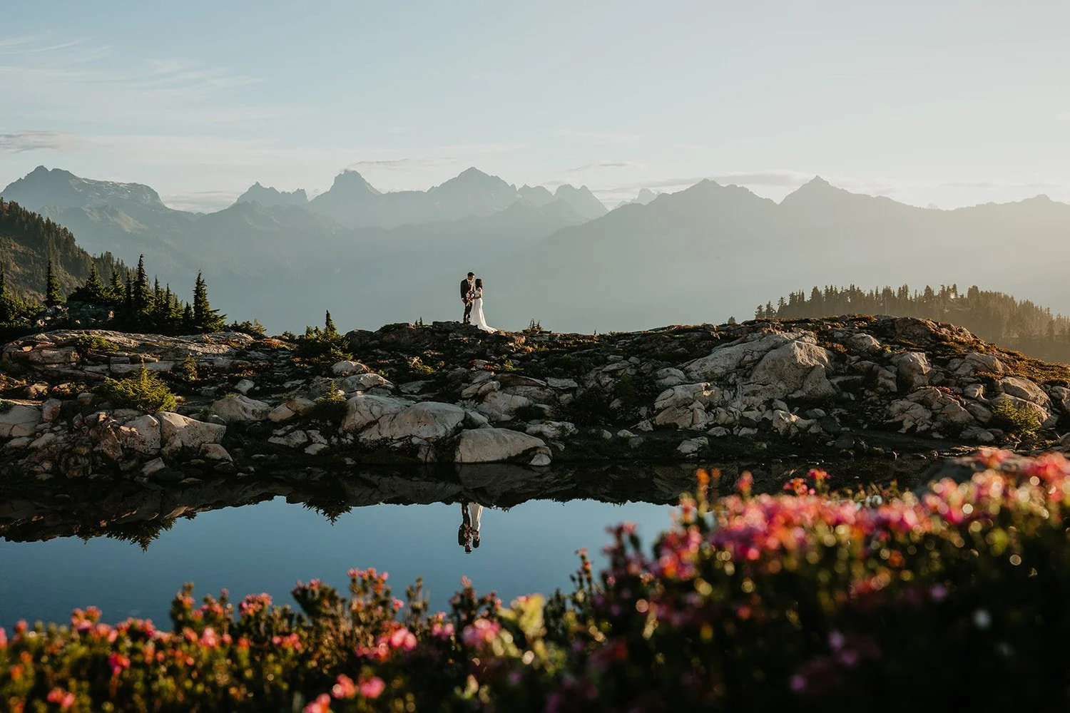 Couple standing on a rocky alpine ridge above a still mountain tarn with wildflowers in the foreground and layered peaks in the distance during golden hour
