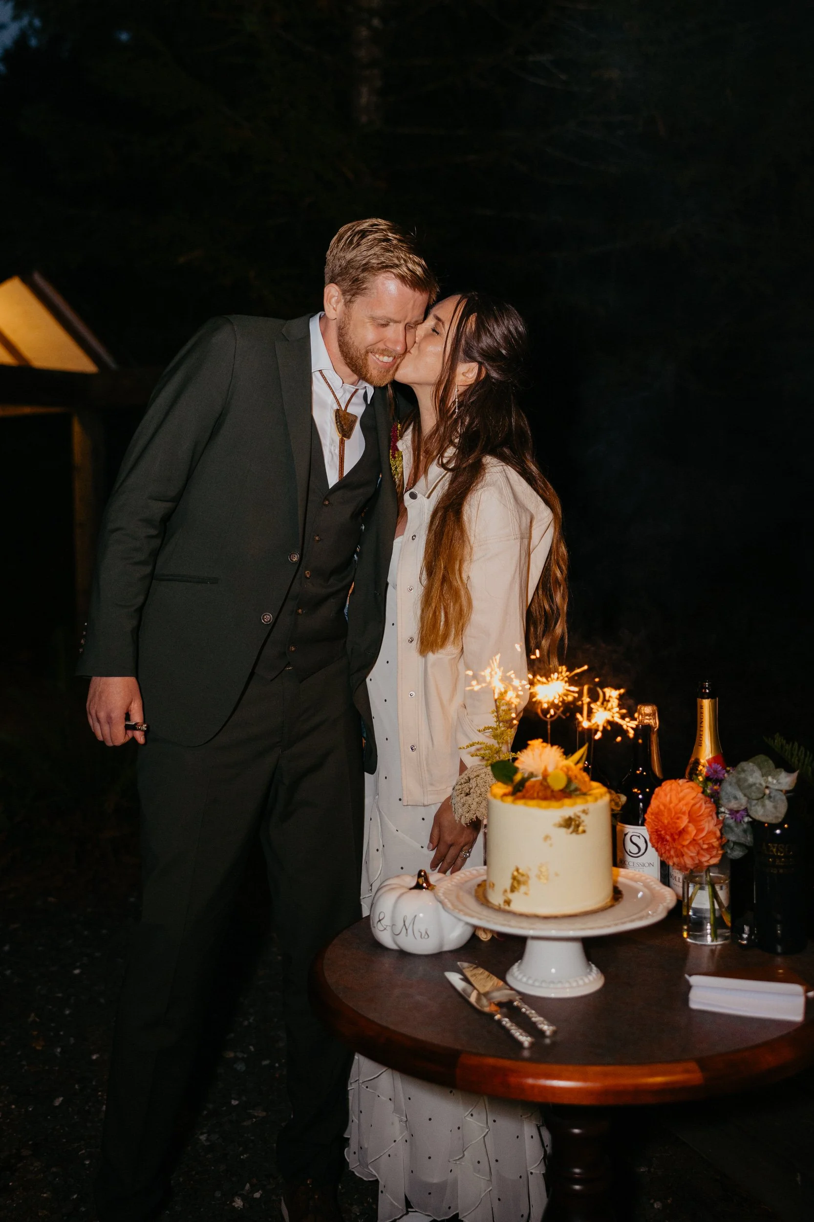 Couple sharing a quiet kiss during their evening elopement celebration in front of their cake with sparklers lit