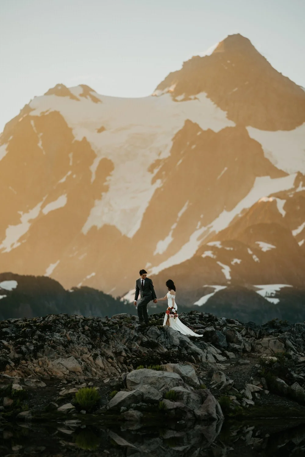 Wide mountain elopement scene with a couple crossing volcanic rock beneath dramatic snow-covered peaks