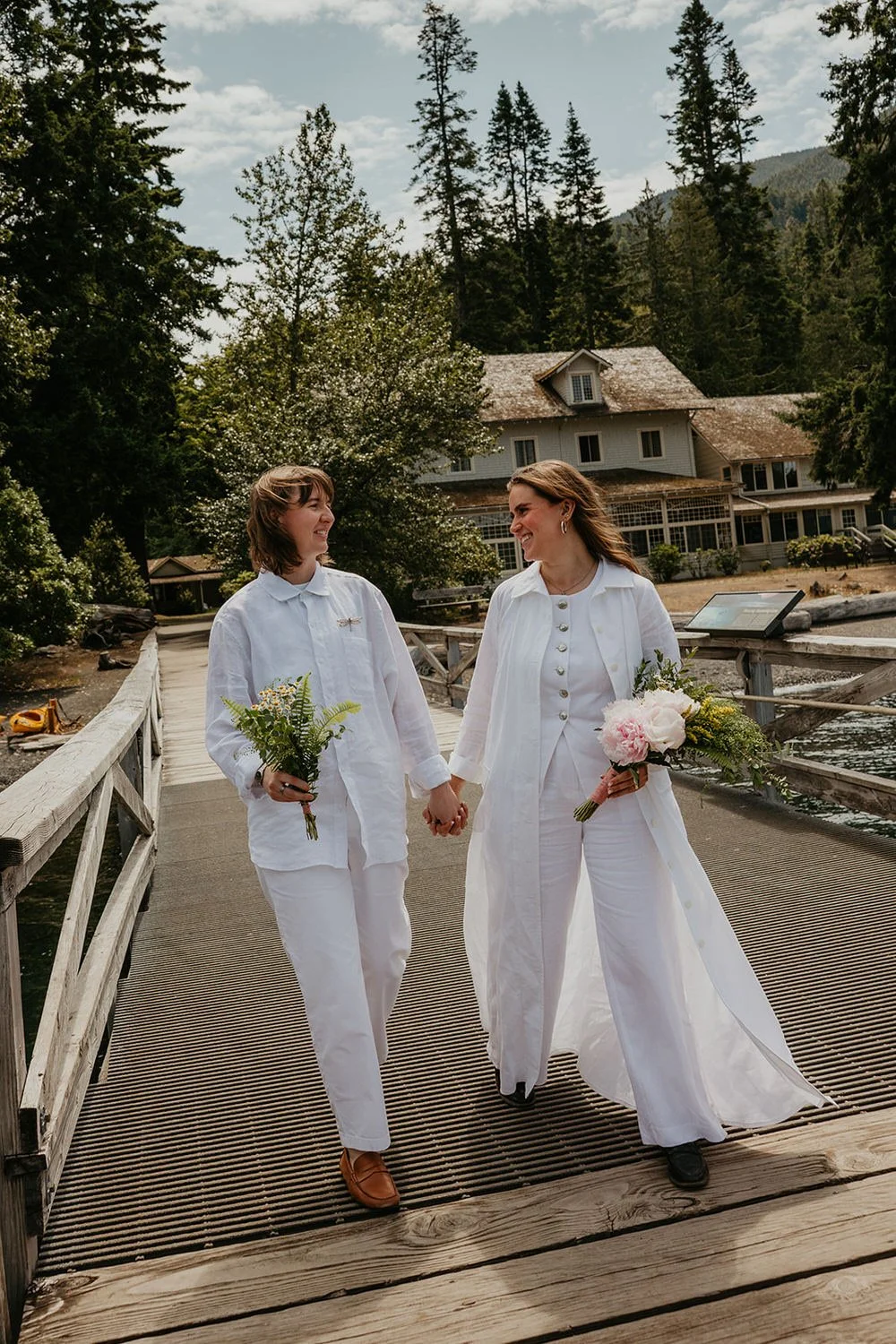 Two brides holding hands while walking across a wooden bridge near a lakeside lodge, smiling at each other during an intimate elopement.