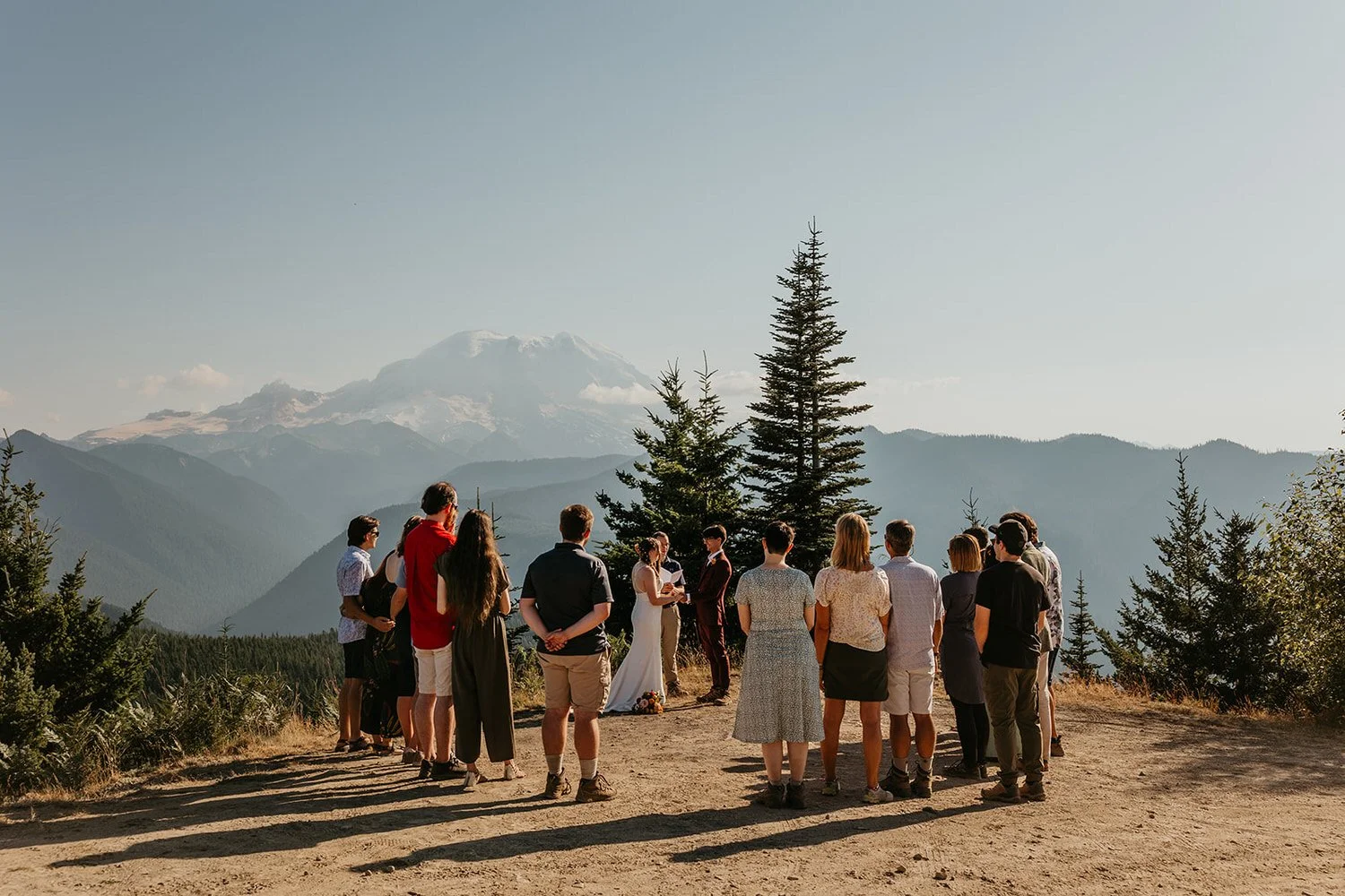 Wedding guests gathered in a semicircle at a mountain overlook during the ceremony, with Mount Baker visible in the hazy distance