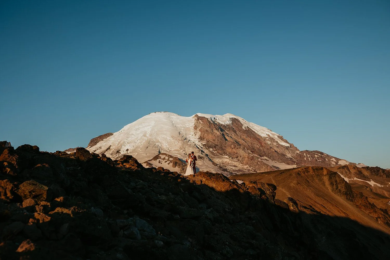 A couple in wedding attire standing on rocky terrain with a snow-capped mountain in the background during sunset.