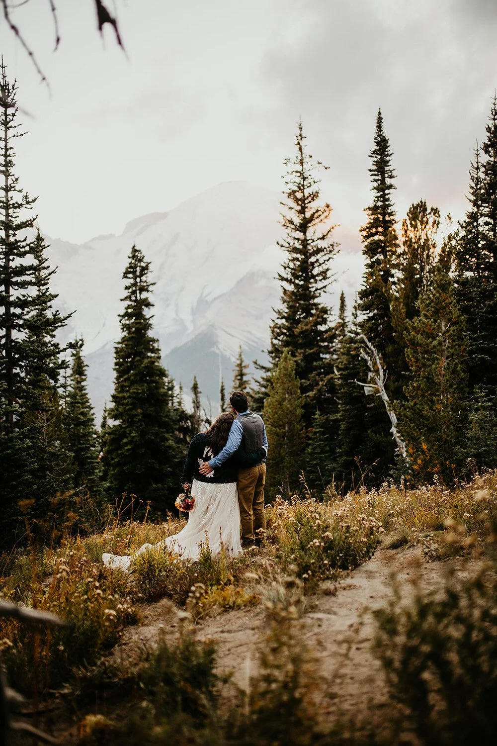 A couple embraces on a narrow trail lined with wildflowers and evergreen trees, facing Mount Rainier in the distance.