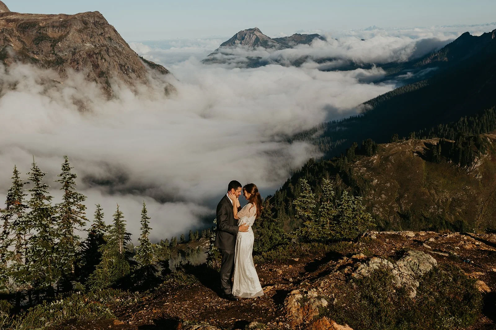 Couple embracing during a North Cascades mountain elopement, standing on an alpine ridge above a sea of clouds with layered mountain peaks in the distance.