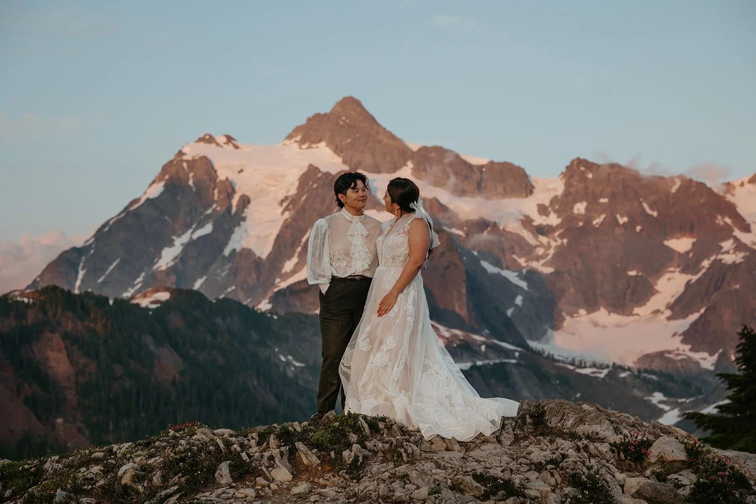 Two women, possibly a bride and same-sex partner, stand close together on a rocky area with snow-capped mountains in the background, during what appears to be sunset or sunrise.