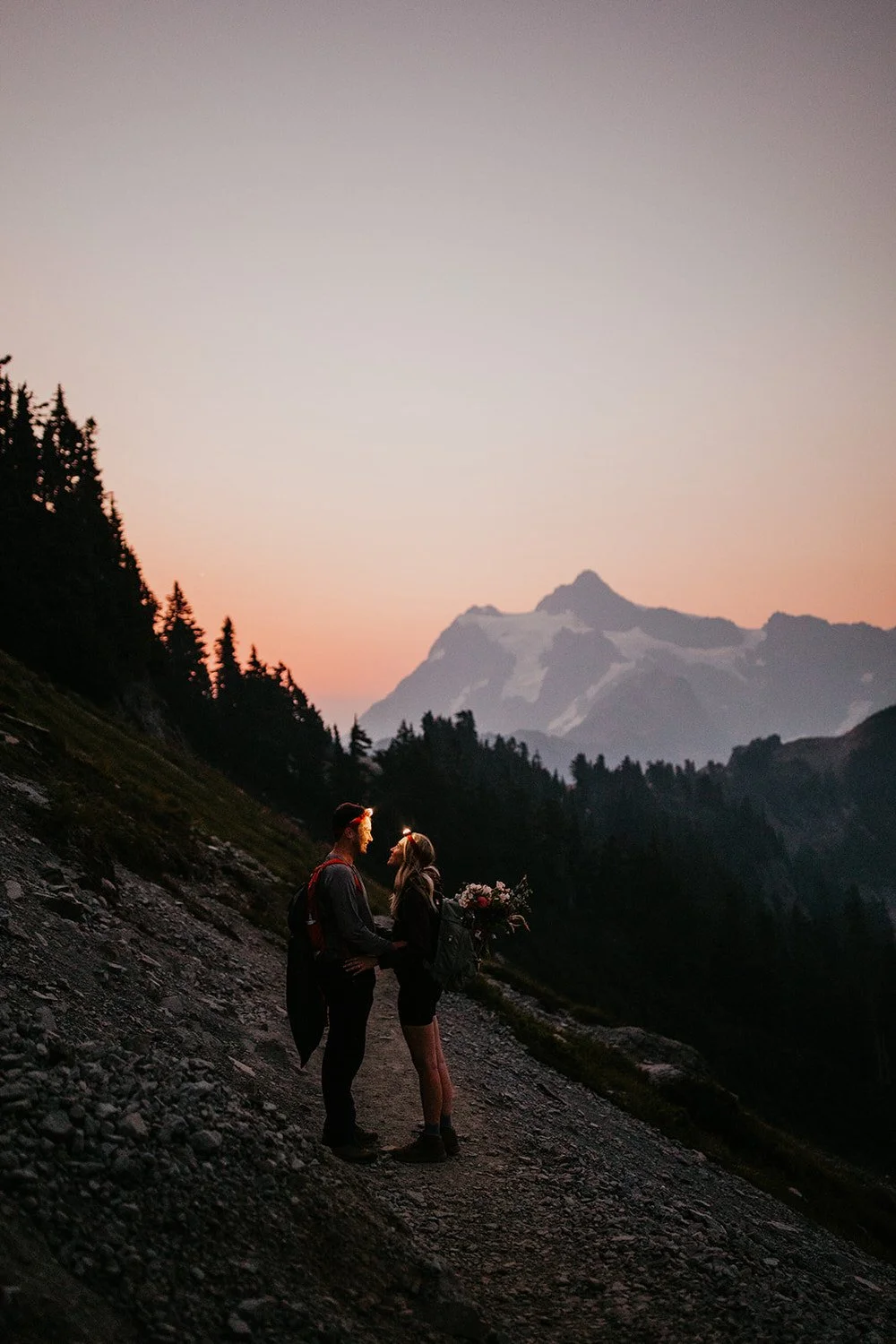 Couple standing face to face on a mountain trail at sunset with headlamps on, surrounded by forested slopes and distant peaks.