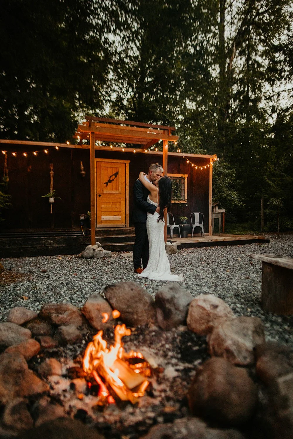 Couple standing together outside a Pacific Northwest cabin at dusk with a glowing fire pit in the foreground during their elopement