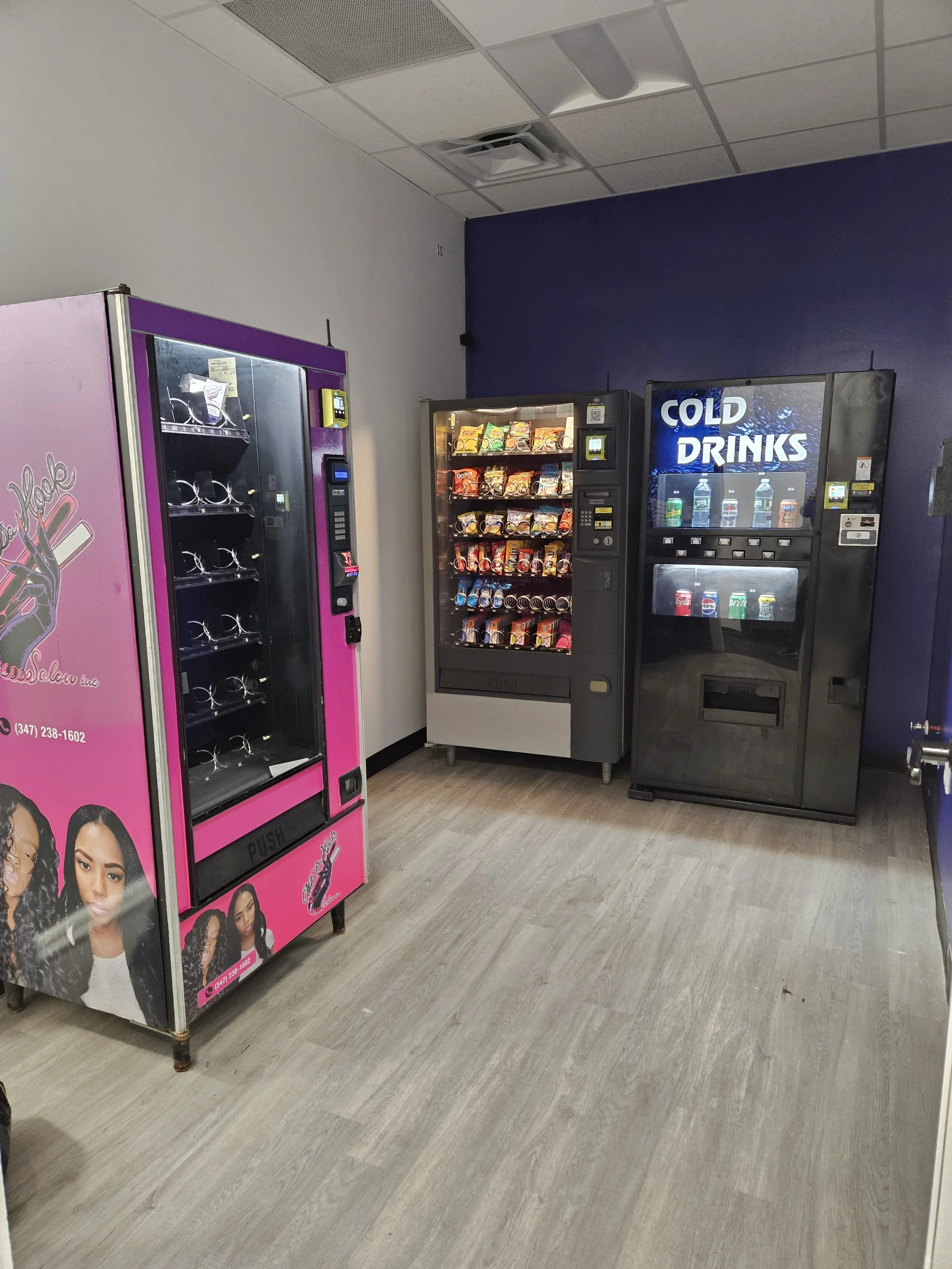 Three vending machines in a room with beige wooden flooring and a dark blue wall. The first machine on the left is pink with images of women on the lower part. The middle machine offers snacks and has a digital payment system. The third machine on the right is a soda vending machine with a digital display that says 'COLD DRINKS' and contains bottled beverages.