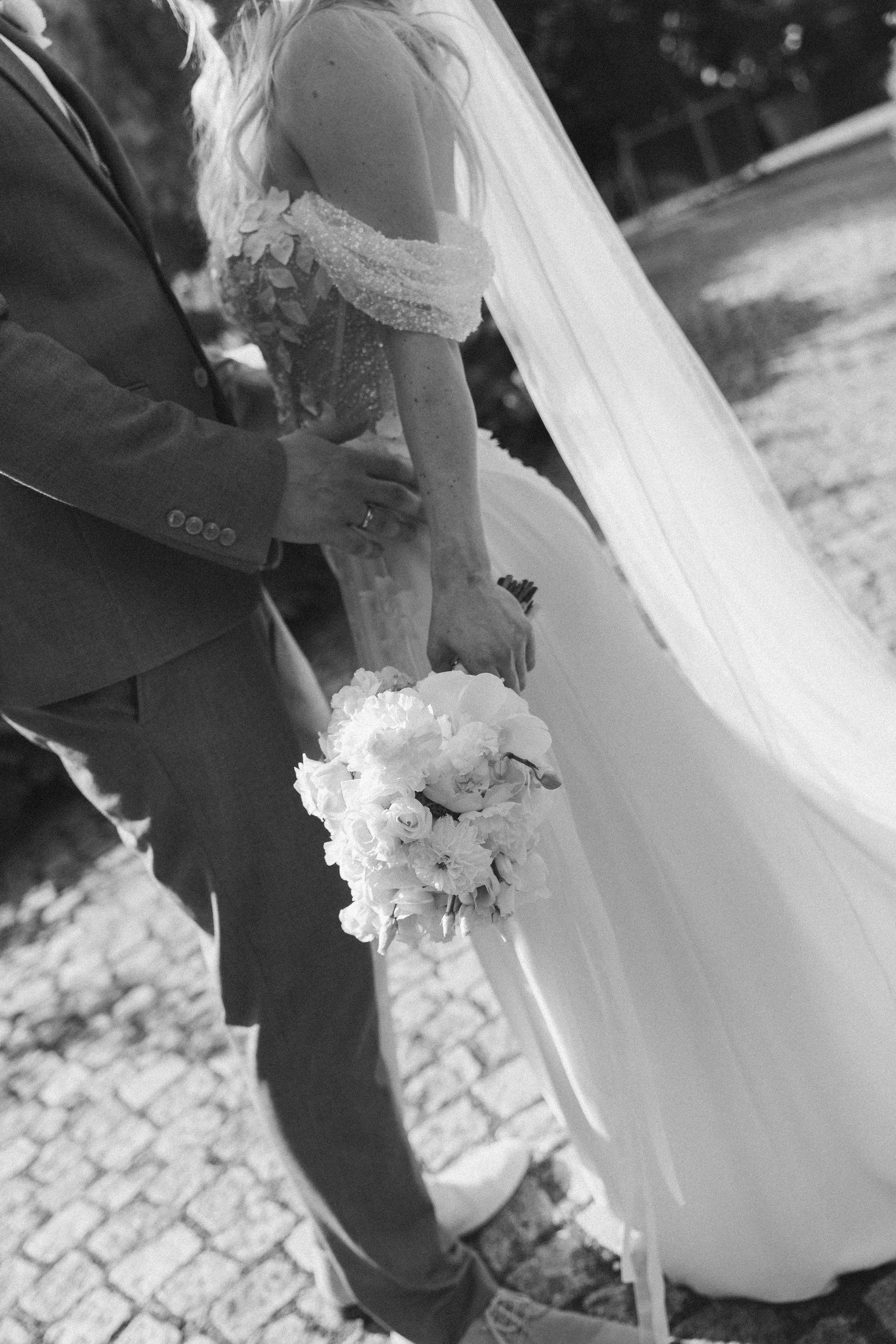 A black and white photo of a bride in a wedding dress holding a bouquet, standing near a person in a suit outdoors on a cobblestone surface.