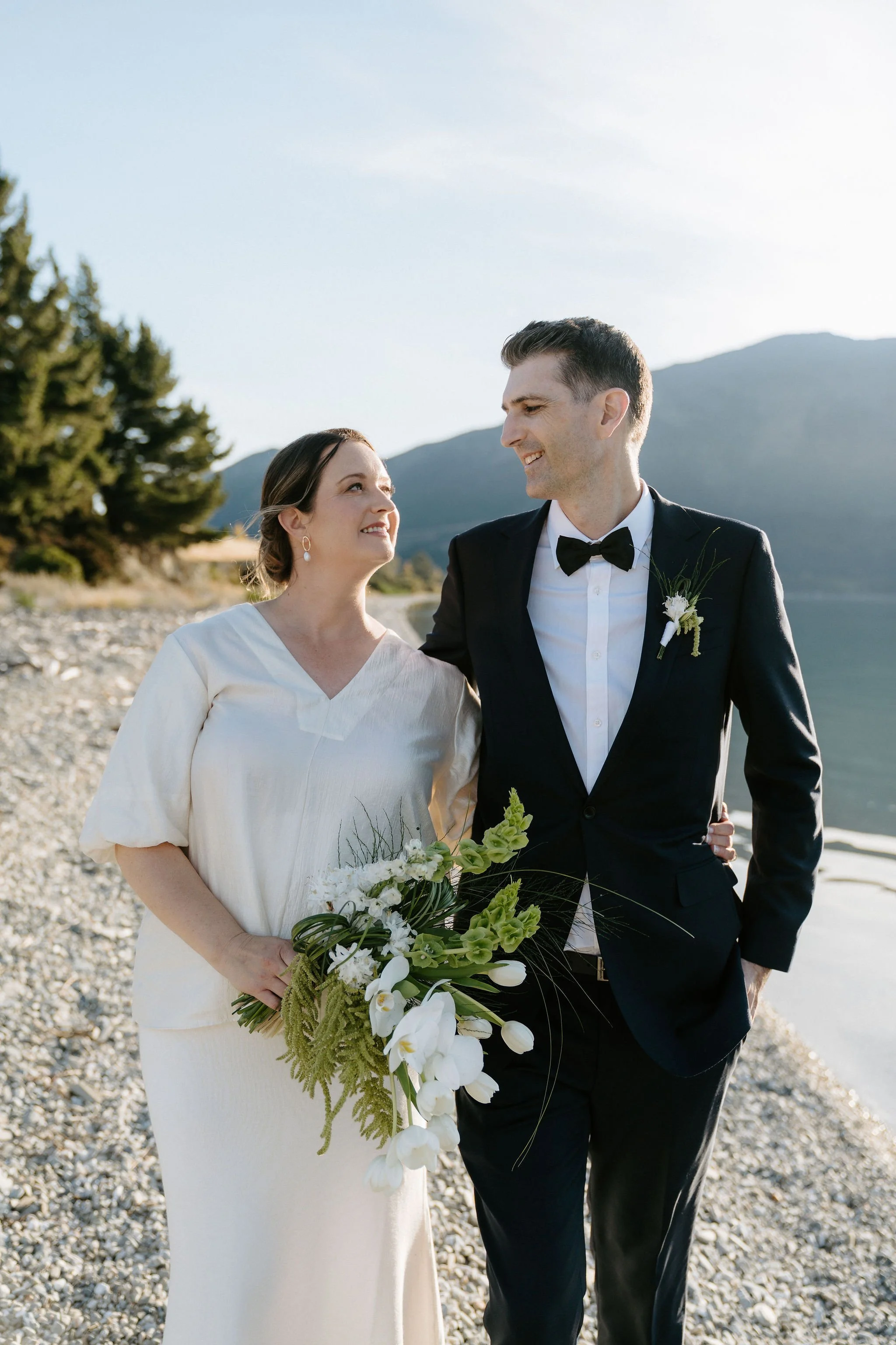 A bride and groom walking on a rocky beach, smiling at each other, with mountains and trees in the background during daylight.