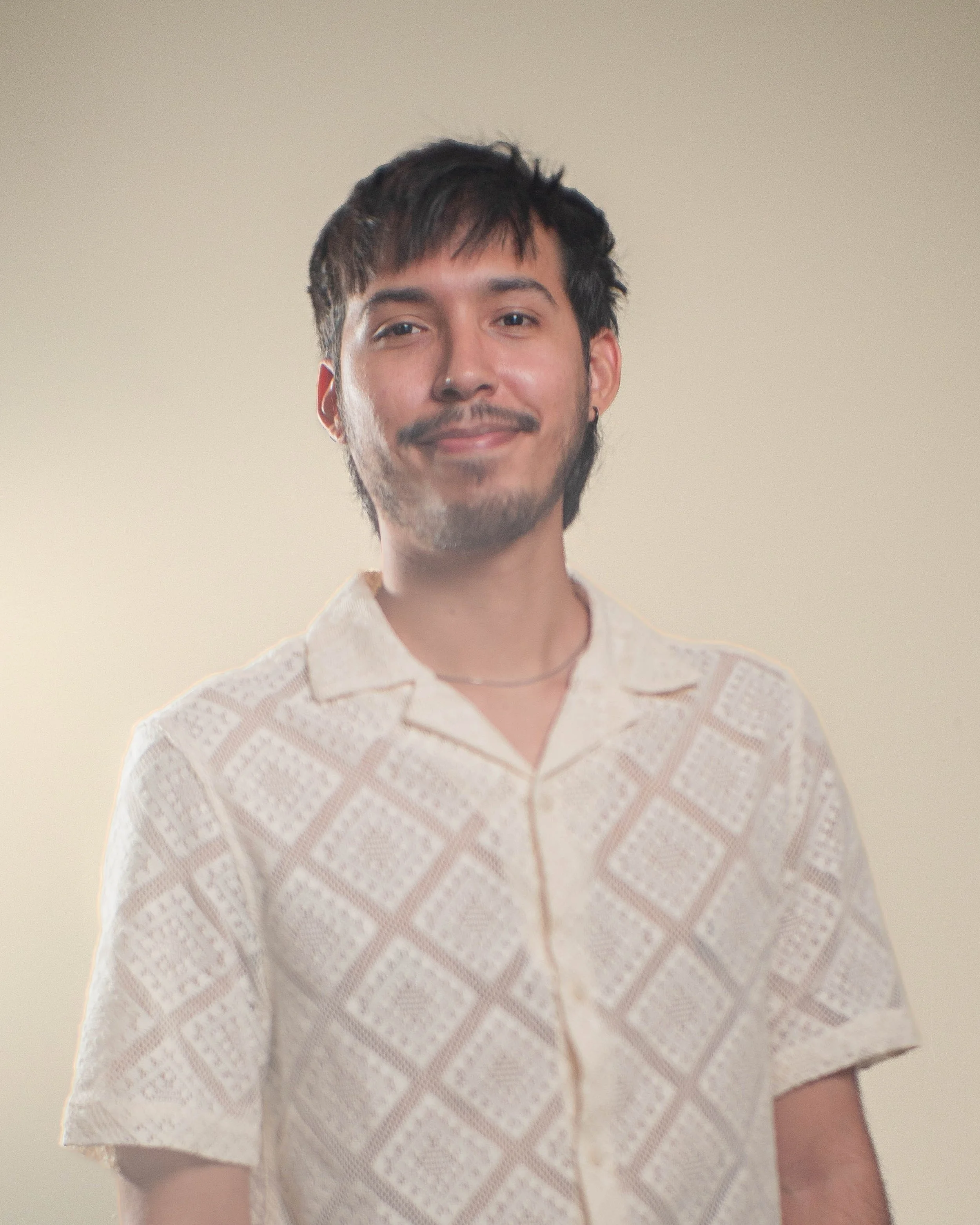 Joven con barba y bigote, cabello oscuro y despeinado, lleva una blusa de manga corta blanca con patrones tejidos, sonríe ligeramente, fondo beige. Enma, Diseñador gráfico.