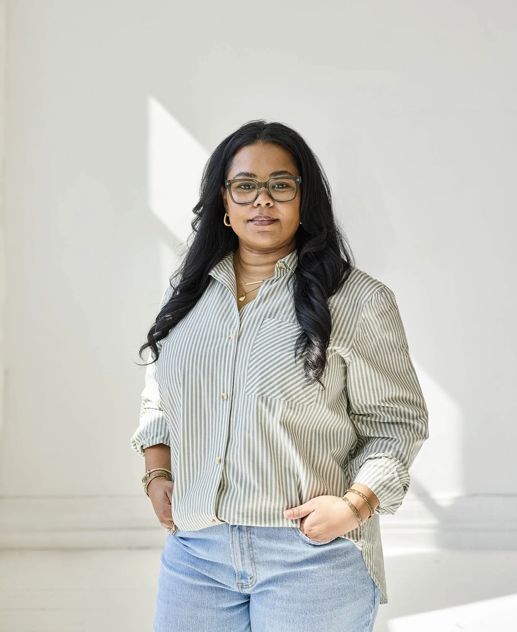 A woman with long black hair, glasses, gold jewelry, wearing a striped button-up shirt and light blue jeans, standing against a plain light-colored wall with sunlight creating a shadow.