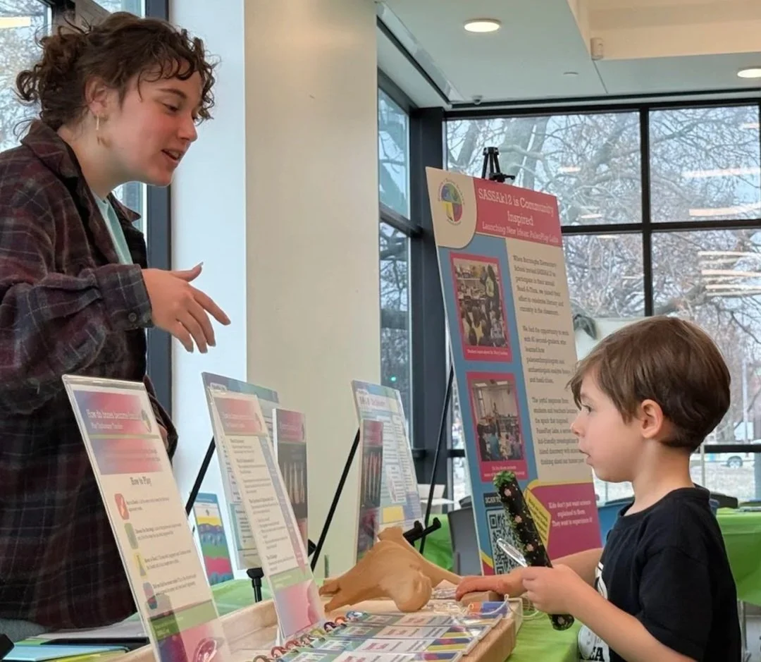 A volunteer stands at a table explaining a fossil activity to a young child, who looks at display boards and holds a tool, with educational materials and signs arranged on stands.