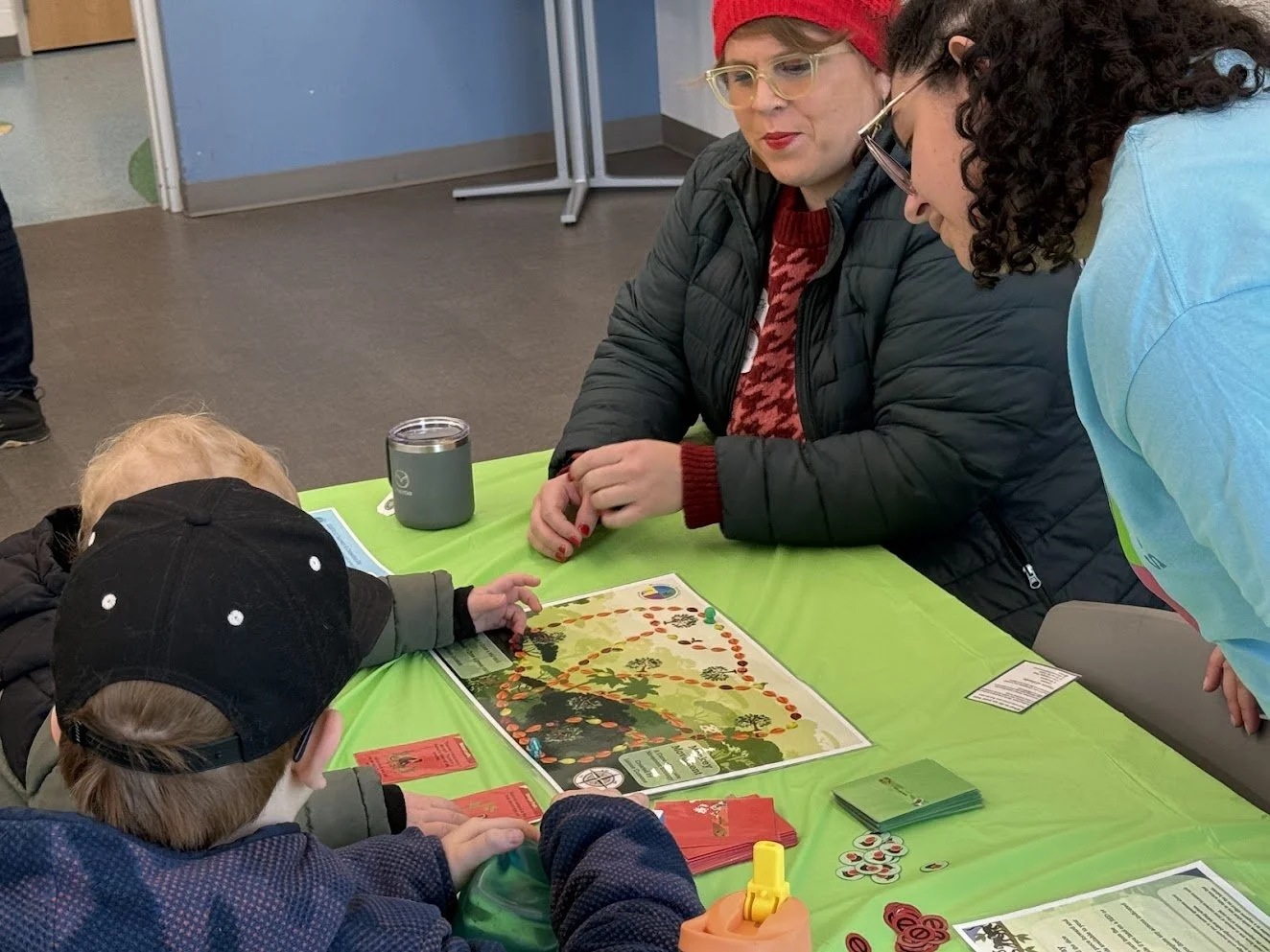 Two adults sit with two young children at a green-covered table, looking at and playing a colorful board game about monkey movement and ecology, with cards and tokens spread around the board.