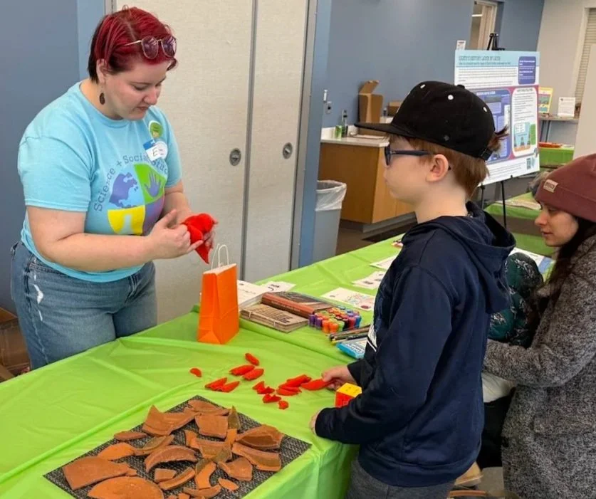 A volunteer stands at a green-covered table guiding two children and a parent as they use clay fragments and red 3D printed elk bone fragments to reconstruct objects. A broken pot puzzle and elk femur puzzle are laid out on the table.