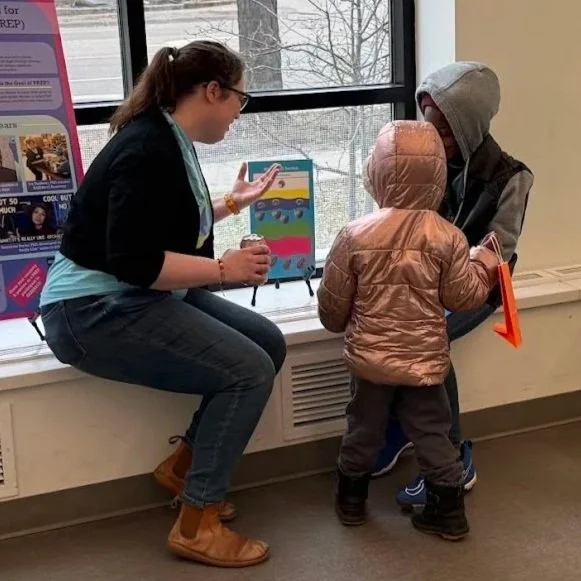 A volunteer sits near a window talking with two children in winter jackets, showing them a colorful educational board while they listen and respond.