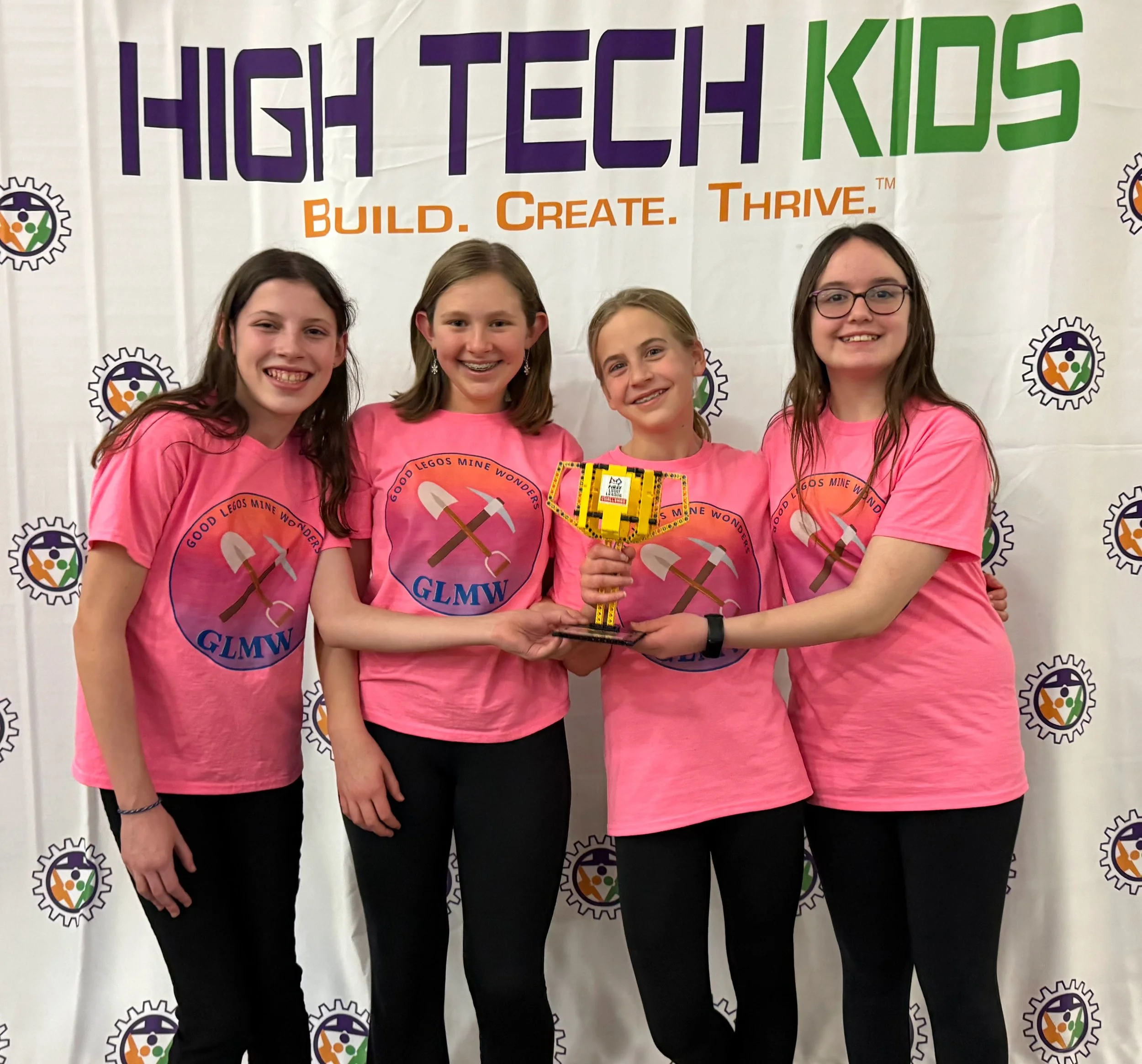 Four members of the Good Legos Mine Wonders FIRST LEGO League team stand together in front of a “High Tech Kids” competition backdrop, holding a trophy. Wearing matching pink team shirts, the students pose closely with arms around one another.