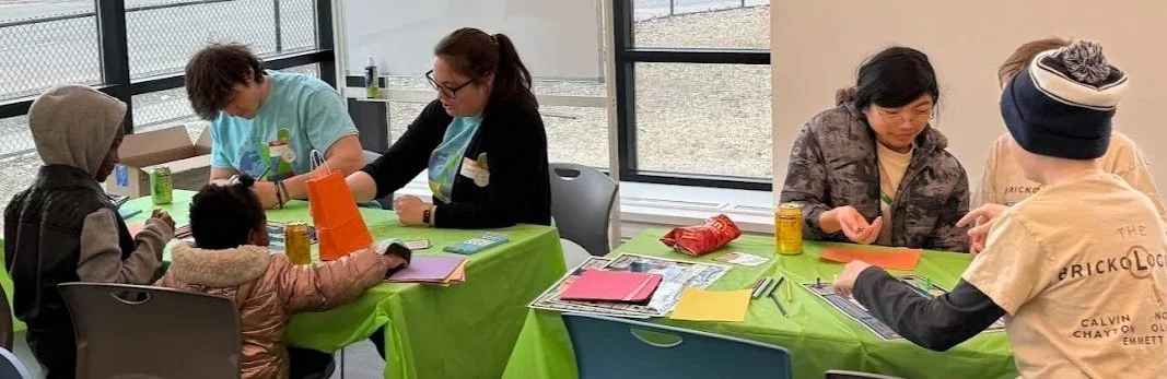 Volunteers and children sit together at two green-covered tables, playing a game about ecological succession, an activity with paper, pencils, and game materials they use to discuss and draw their outcomes.