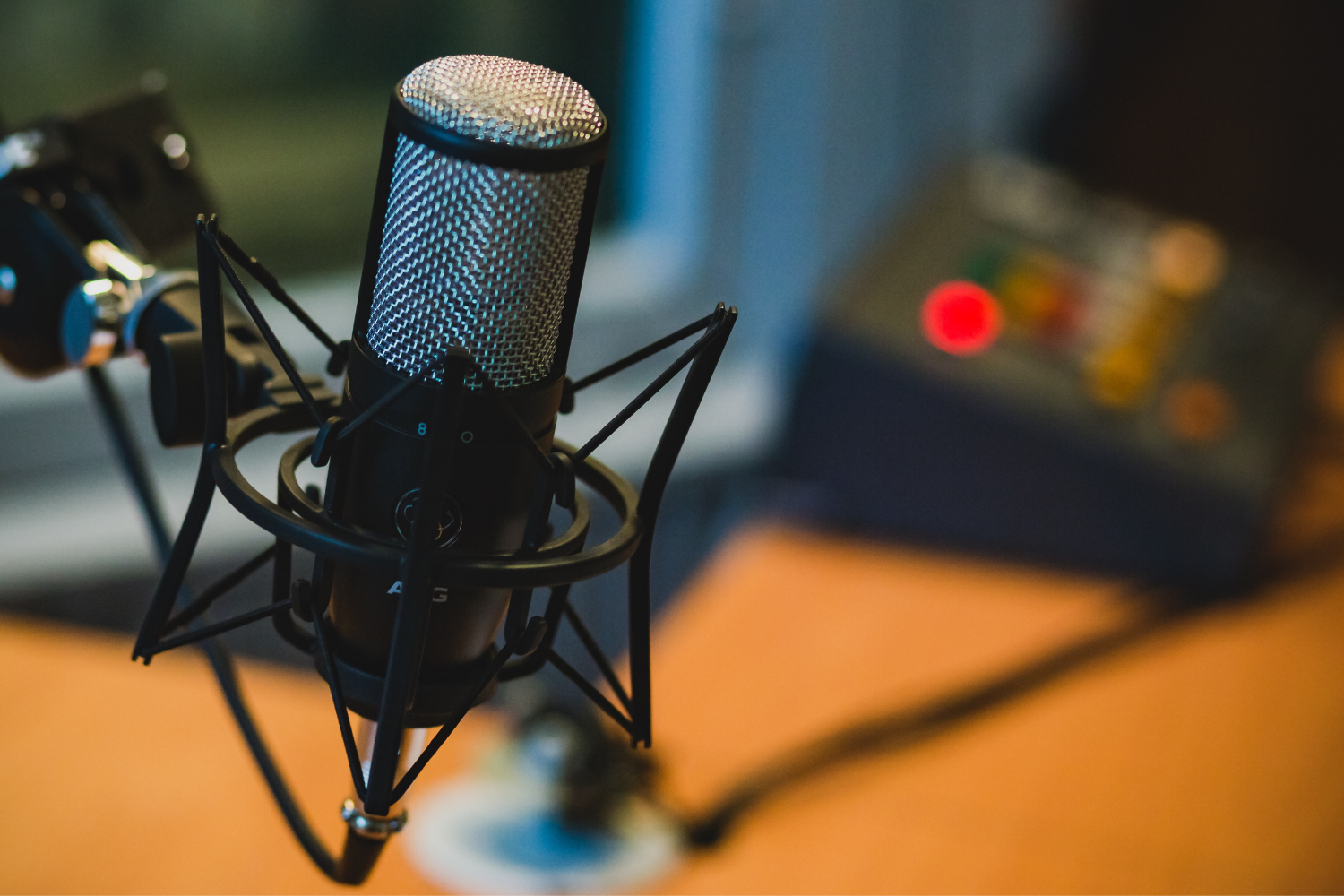 Close-up of a studio microphone with a pop filter mounted on a stand, set on a wooden desk, with a blurred background of audio equipment.