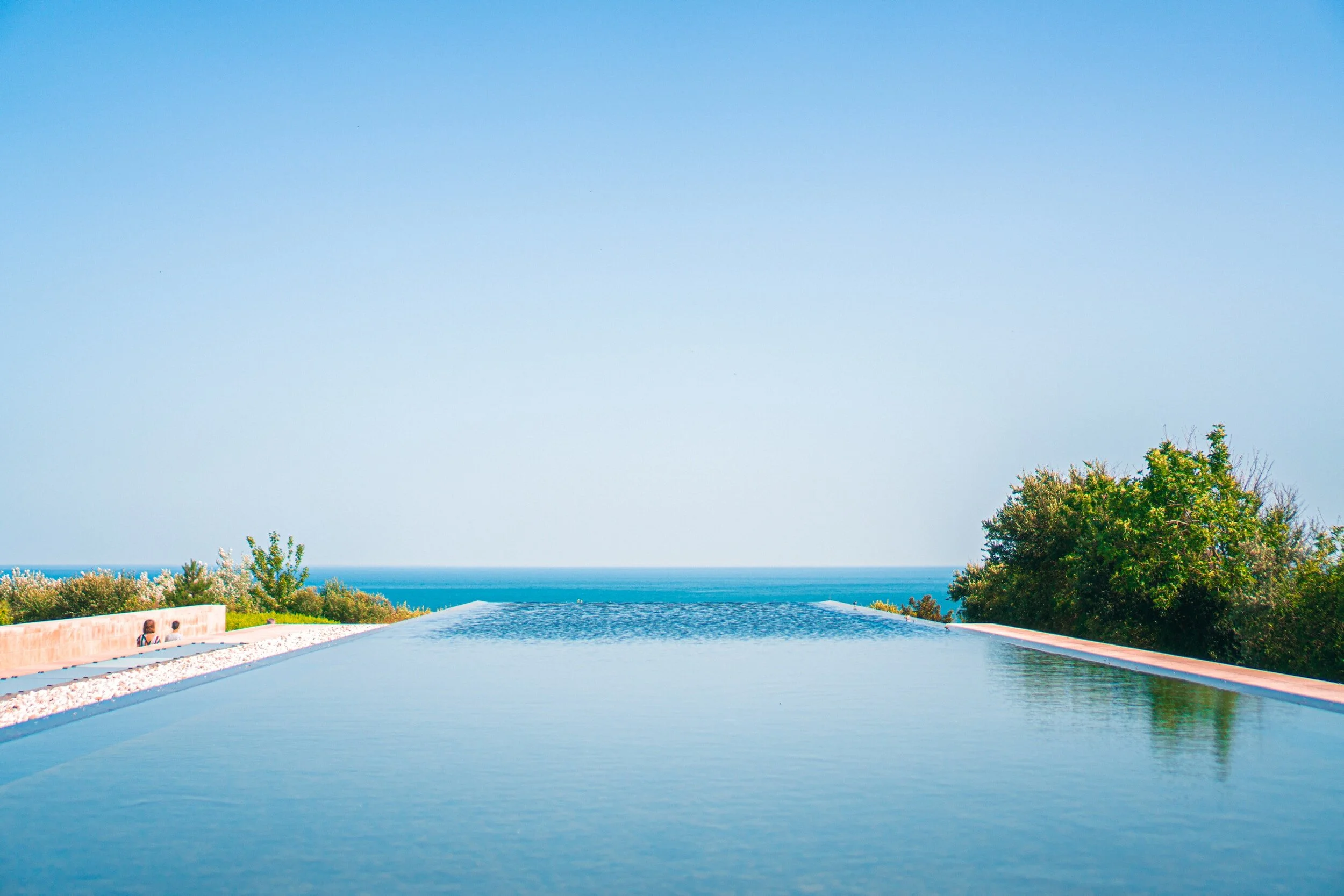 Infinity pool overlooking the ocean, surrounded by greenery, under a clear blue sky.