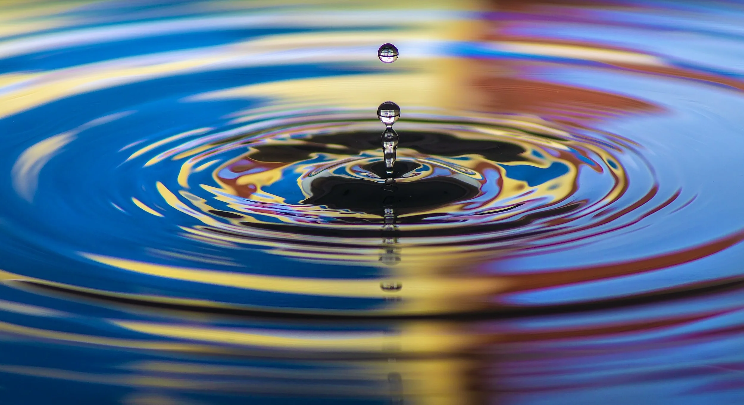 Close-up of a water droplet falling into colorful water, creating ripples and reflections with vibrant colors.