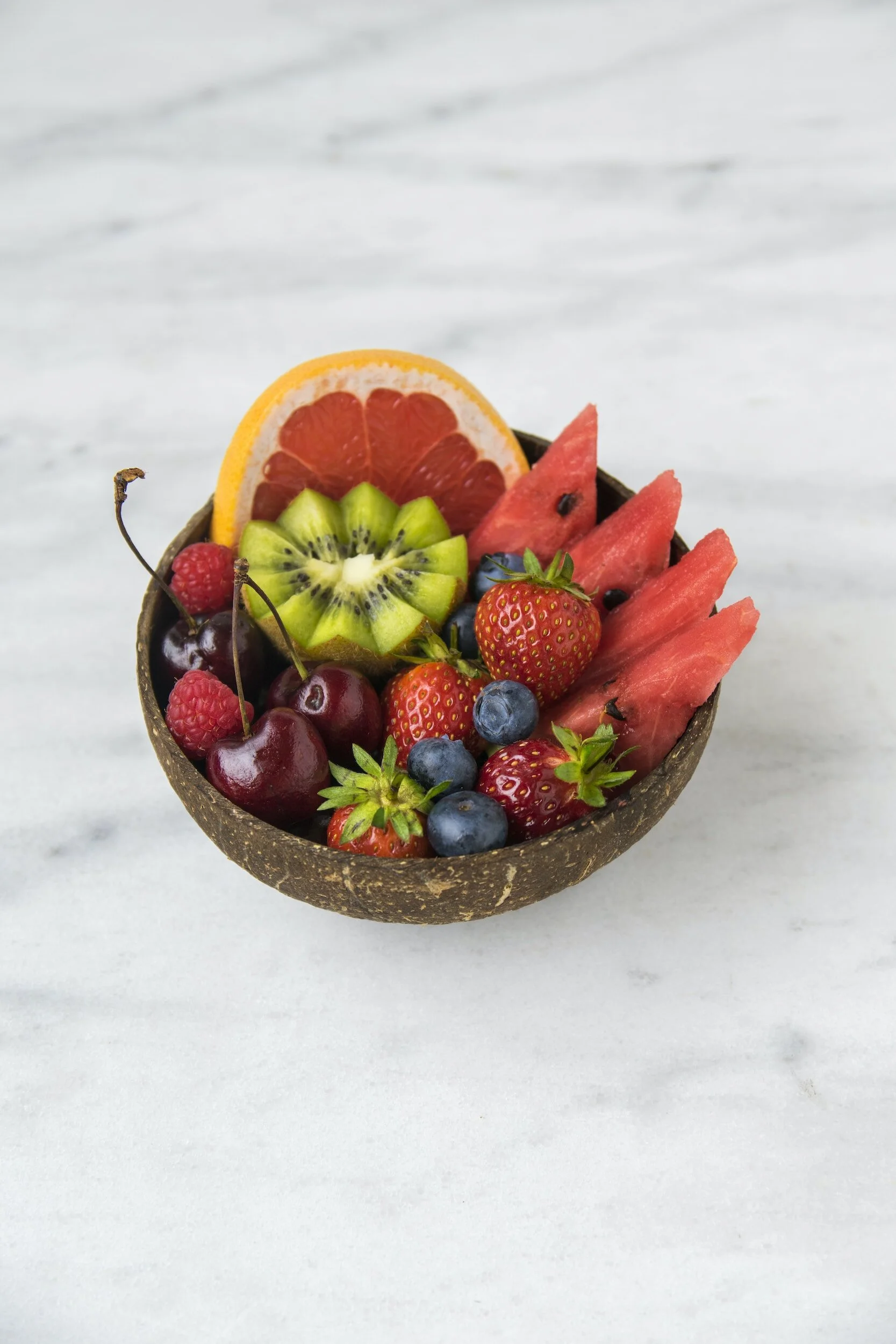 A bowl of assorted fresh fruits including watermelon, kiwi, grapefruit, strawberries, blueberries, cherries, and raspberries on a marble surface.