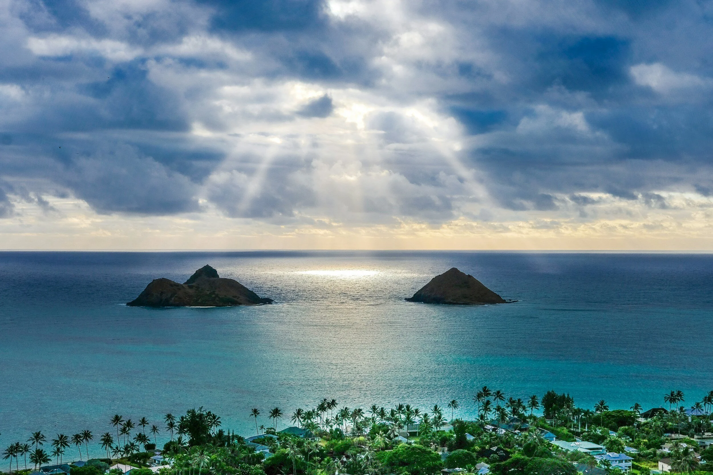 View of two small islands in the ocean with sunlight breaking through cloudy sky and a tropical beachfront in the foreground.