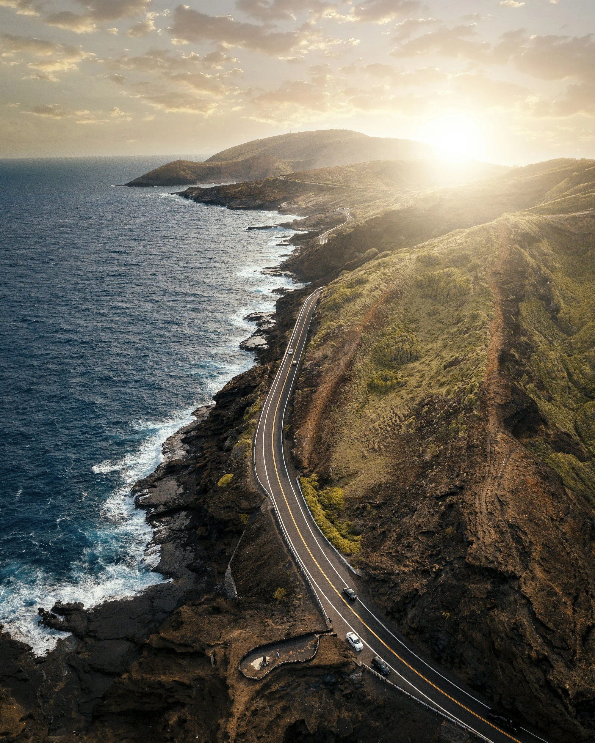 Aerial view of a coastal road winding along a rugged cliffside with ocean waves crashing below and a bright sunset on the horizon.