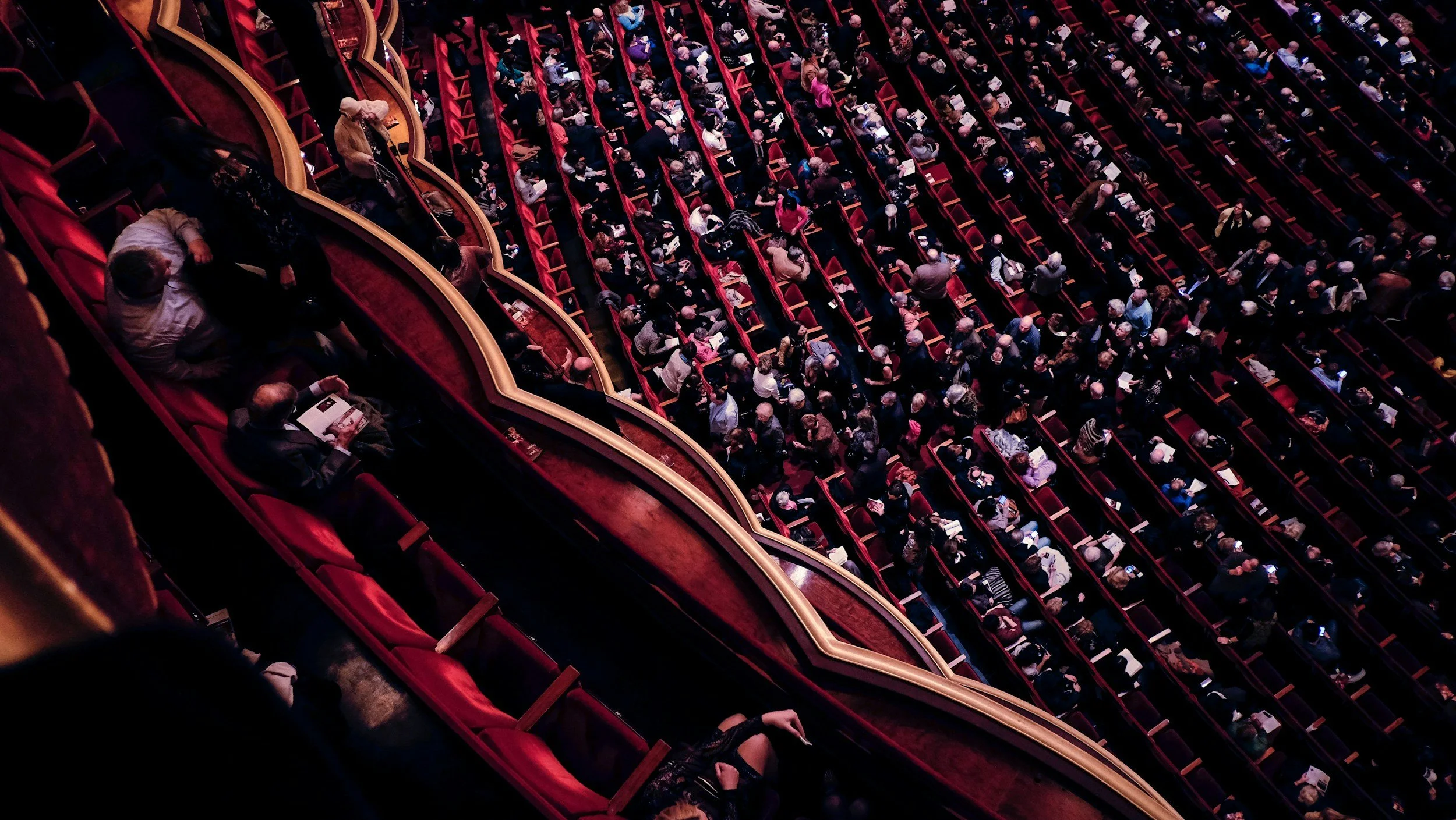 High angle view of audience seated in a theater with red seats and ornate gold railings.
