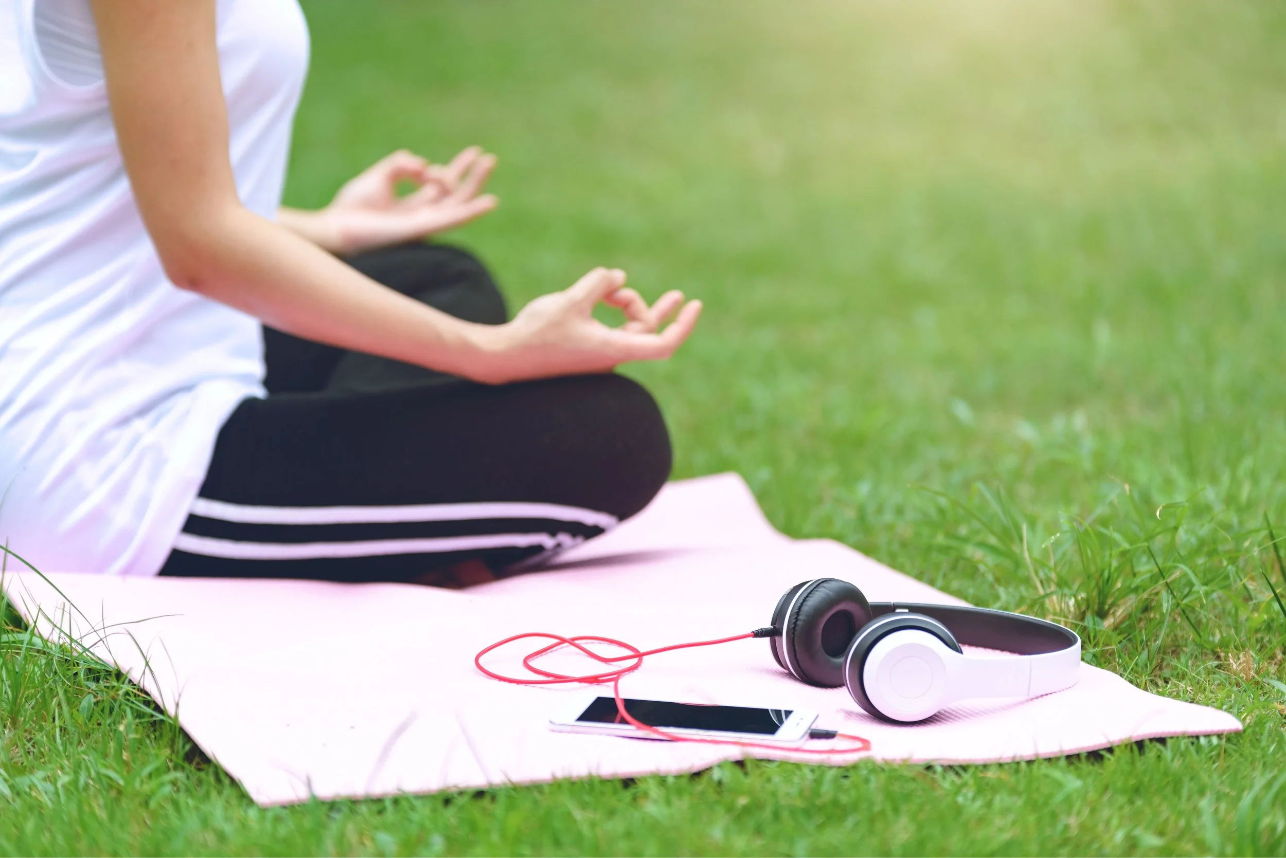 Person sitting cross-legged on a yoga mat outdoors, with headphones and a smartphone nearby on the grass.