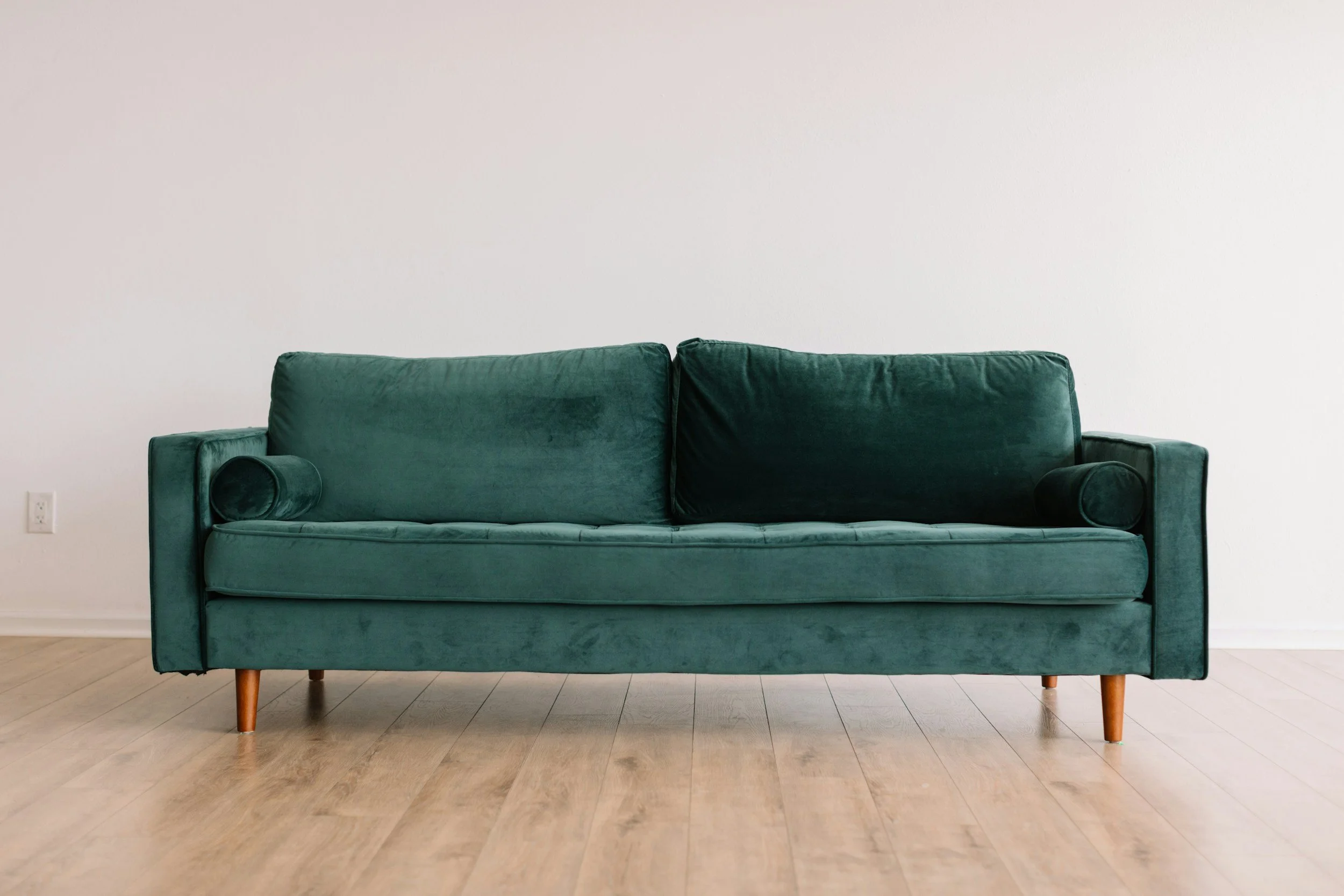 A green velvet sofa with wooden legs placed against a plain white wall in a room with light wood flooring.