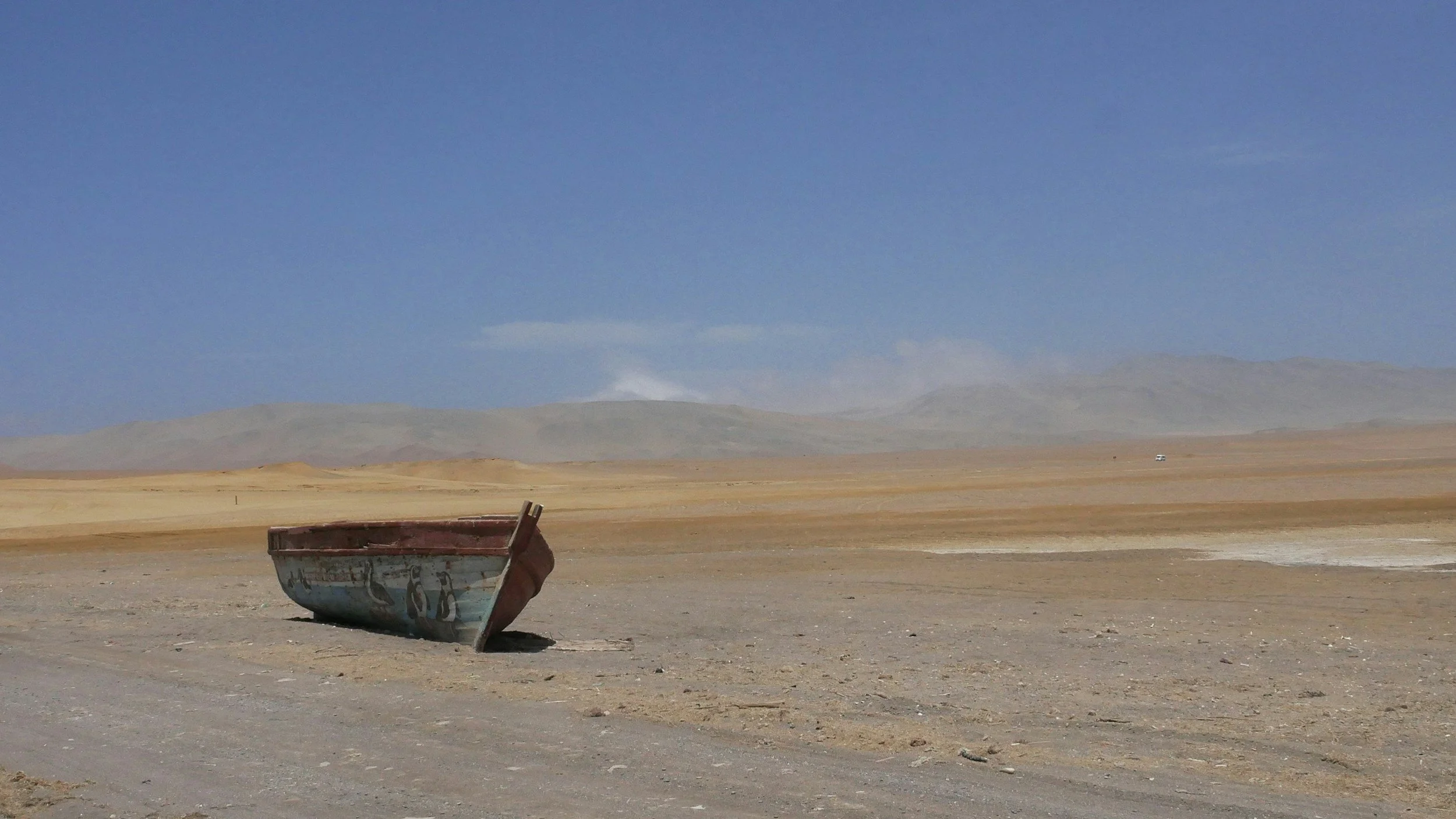 An abandoned boat resting on dusty ground in a barren desert landscape with distant hills and a clear blue sky.