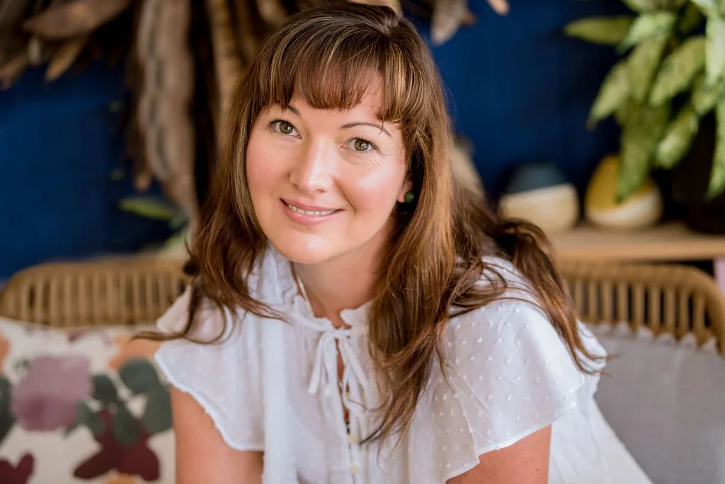 A woman with long brown hair and a smile, wearing a white blouse with a bow, sitting on a sofa in a room decorated with plants and cushions.