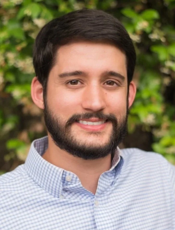 A young man with dark hair and a beard smiling outdoors, wearing a light checkered button-up shirt, with blurred green foliage in the background.