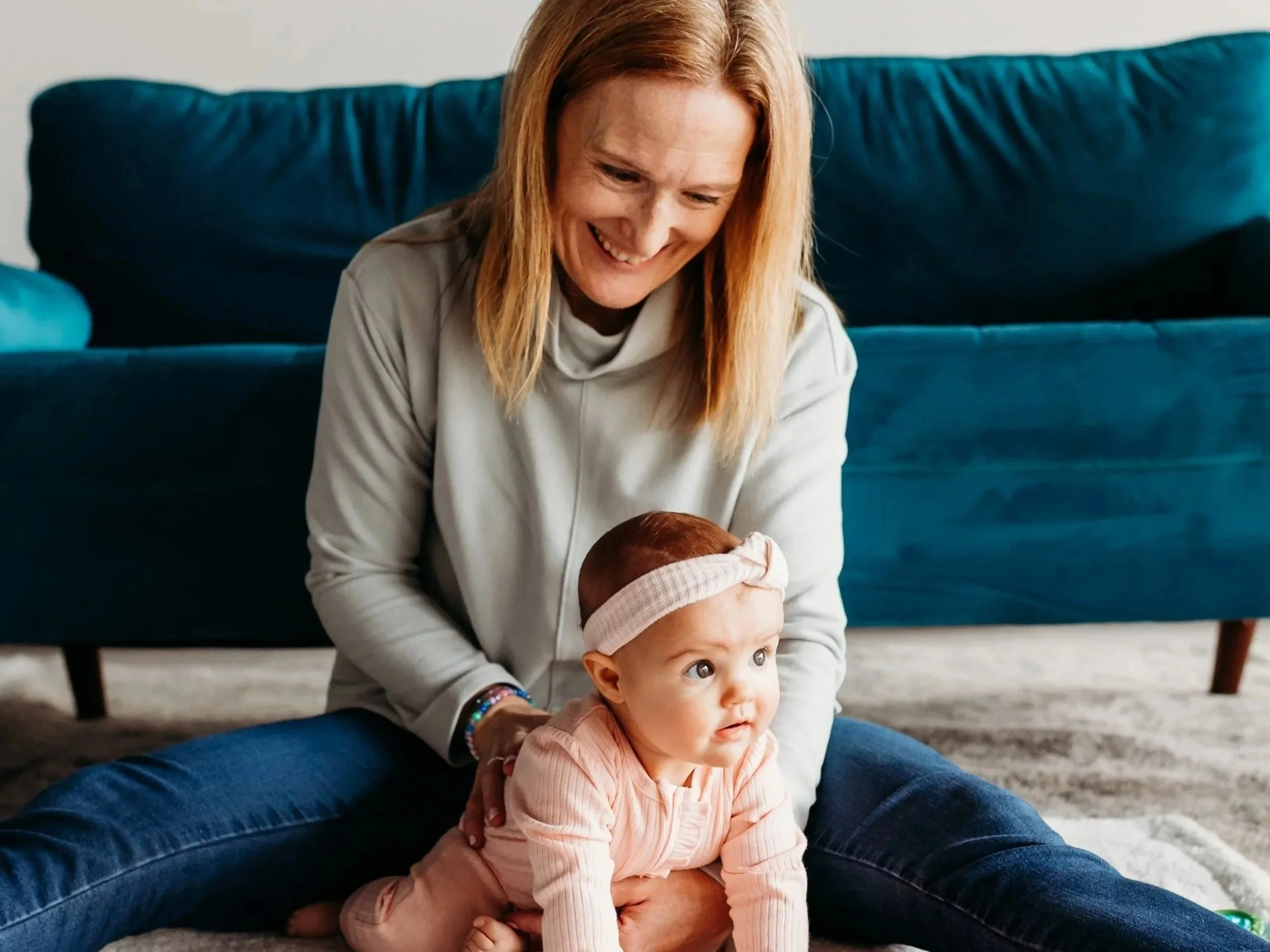 A woman with red hair smiling and playing with a baby girl sitting on the floor in front of her, in a living room with a blue velvet couch in the background.