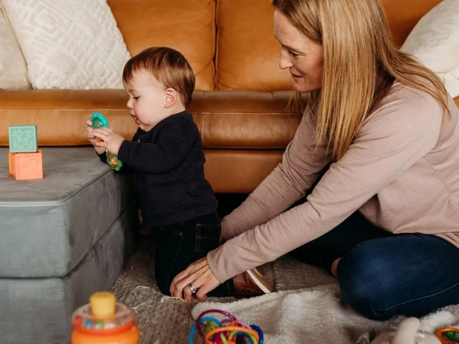 A woman helping a young child stand on the floor while playing with toys in a cozy living room.